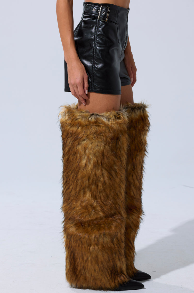 Someone stands against a plain light background wearing the BUILD ME UP FAUX LEATHER SHORT in black, featuring a high-rise fit with buckle detail, paired with tall brown furry boots that reach above the knees. 5