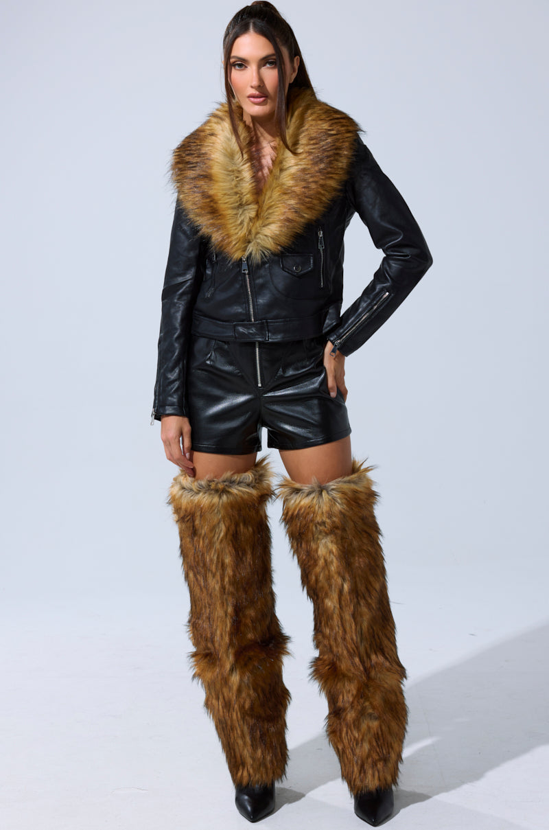A woman faces forward, wearing the BUILD ME UP FAUX LEATHER SHORT, a black leather jacket with a large faux fur collar, and matching tall faux fur leg warmers. Her long hair is styled straight. 7