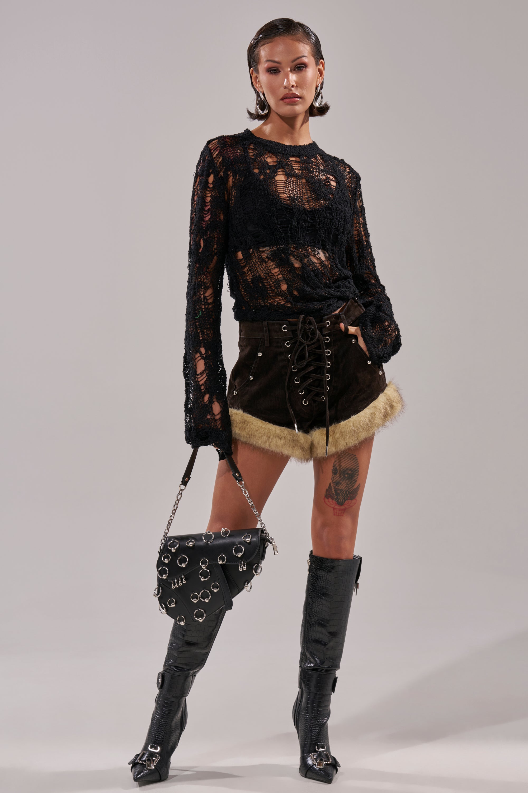 A woman poses confidently in a sheer black lace top with a black bra, ANGEL OF MINE DENIM LACE UP SHORT, knee-high black boots, and a sleek handbag with silver rings. Her hair is slicked back and her makeup is bold.