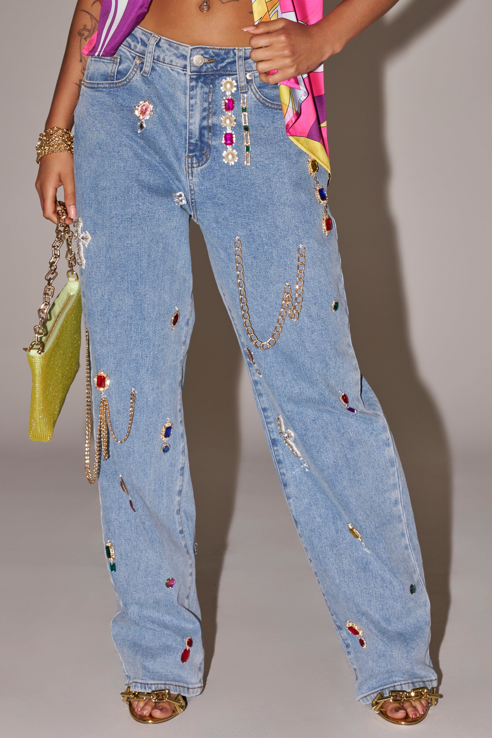 A person wearing FEELING THE DIFFERENCE EMBELLISHED RELAXED DENIM jeans with colorful rhinestones and gold chains, paired with a yellow sparkly handbag and gold sandals. 0
