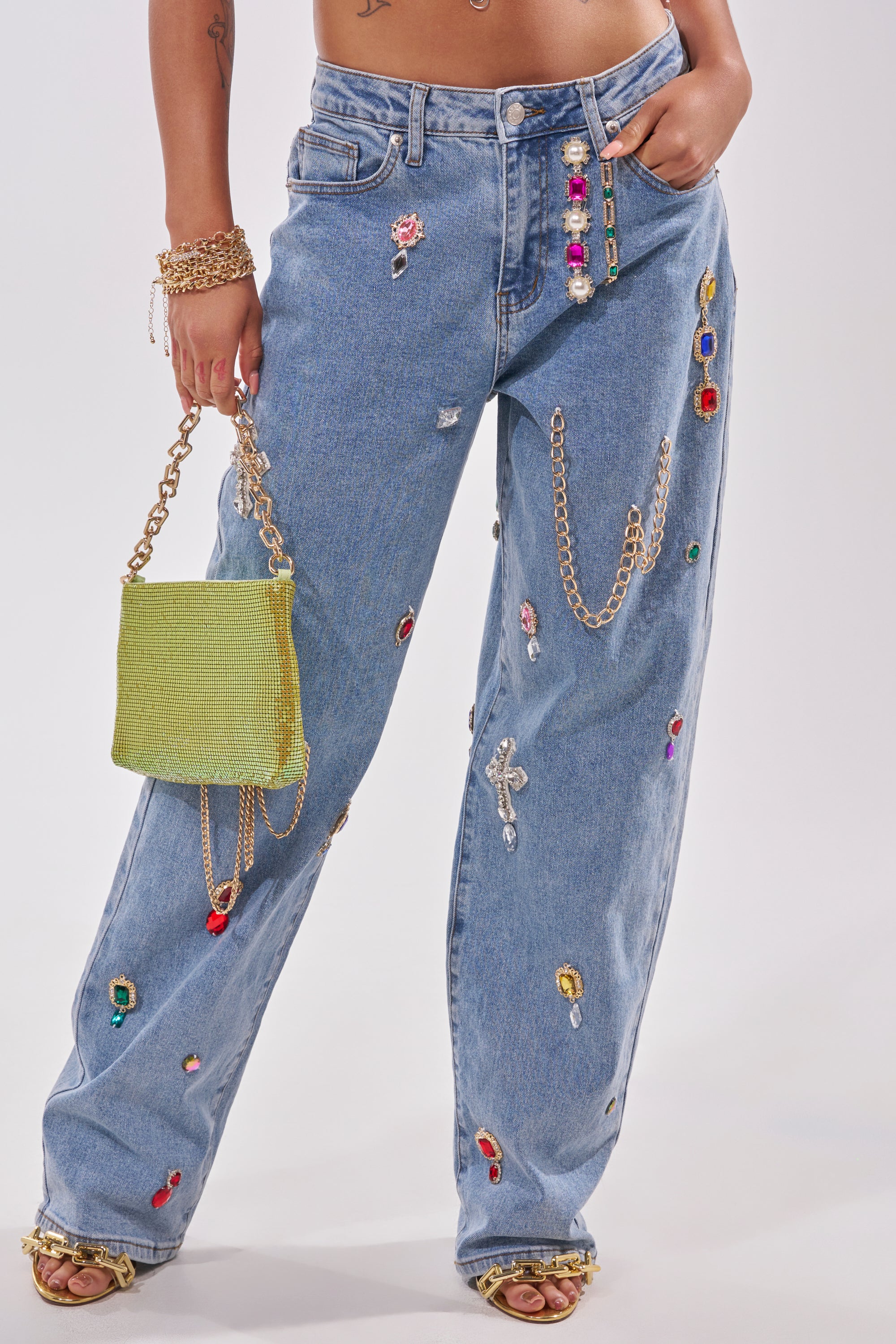 A person wears the FEELING THE DIFFERENCE EMBELLISHED RELAXED DENIM—high-rise blue jeans with colorful gems and gold chains—paired with a shiny green handbag, gold sandals, and bracelets against a plain light background. 2