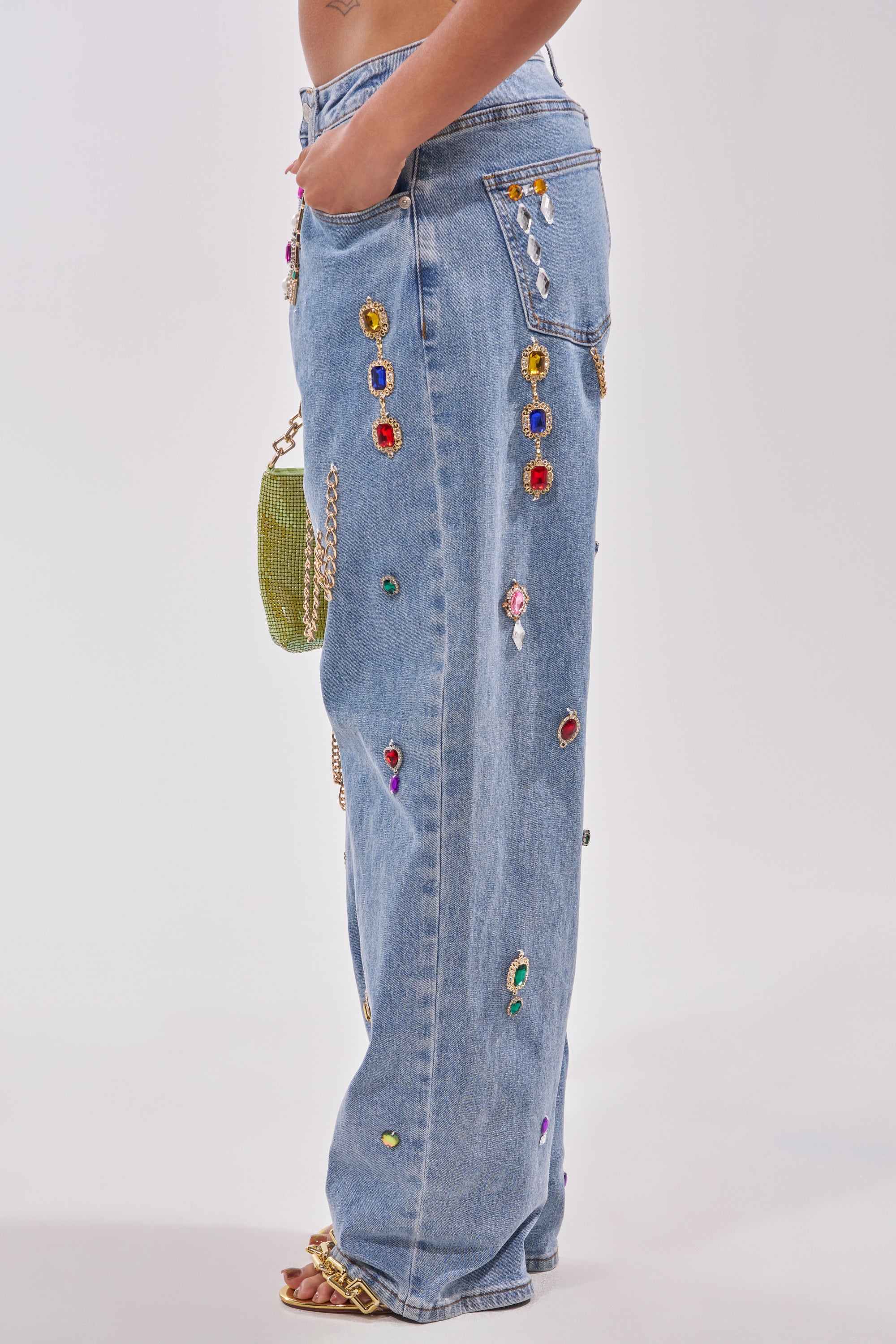 A person wearing FEELING THE DIFFERENCE EMBELLISHED RELAXED DENIM in light blue with colorful jeweled details and a gold chain purse, shown from the side against a plain background. 3