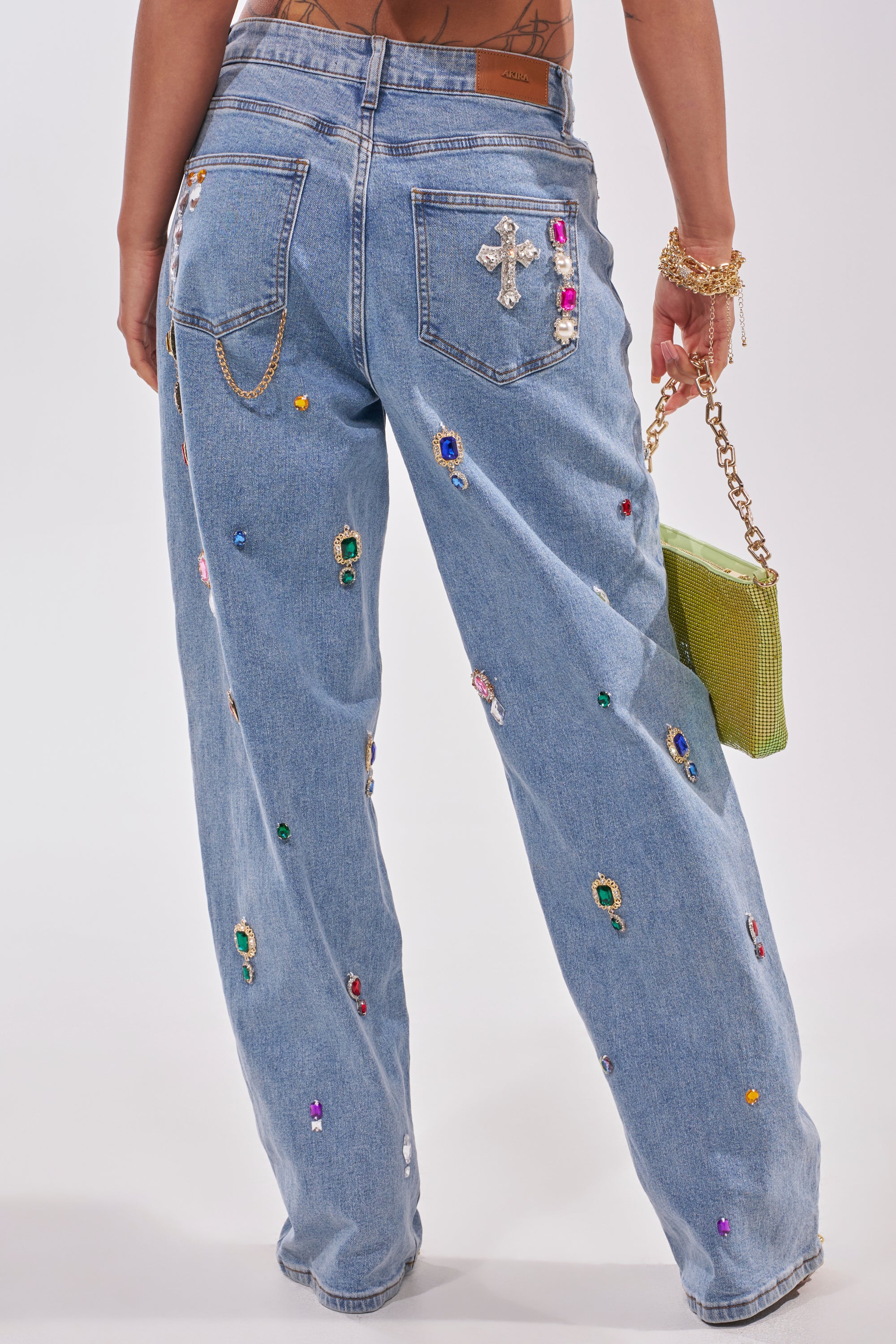 Wearing FEELING THE DIFFERENCE EMBELLISHED RELAXED DENIM—light blue jeans with colorful gemstones and a jeweled cross on the back pocket—a person holds a small green handbag and gold bracelets. Photo is taken from behind. 4