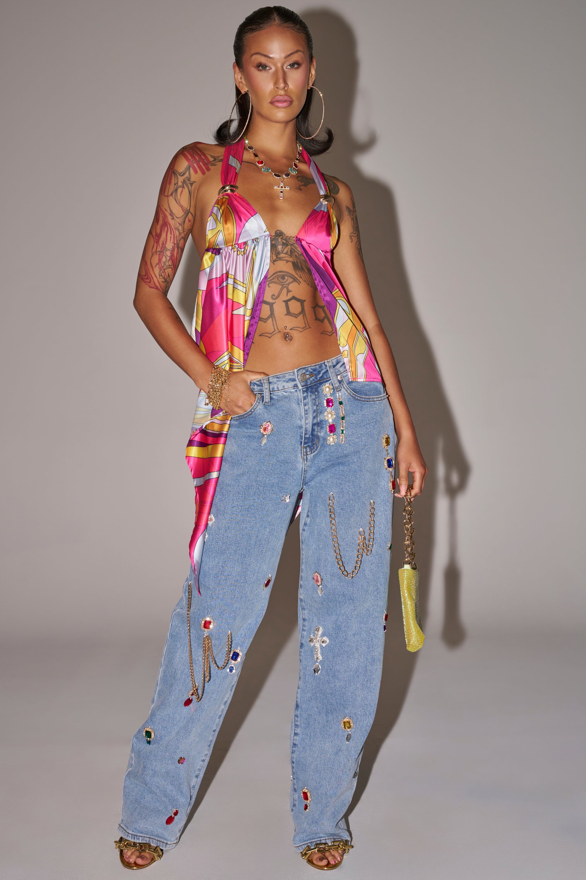 A woman poses confidently in a colorful scarf top, FEELING THE DIFFERENCE EMBELLISHED RELAXED DENIM, and heeled sandals, accessorizing with hoop earrings, layered necklaces, and a small yellow purse with a chain strap. 5