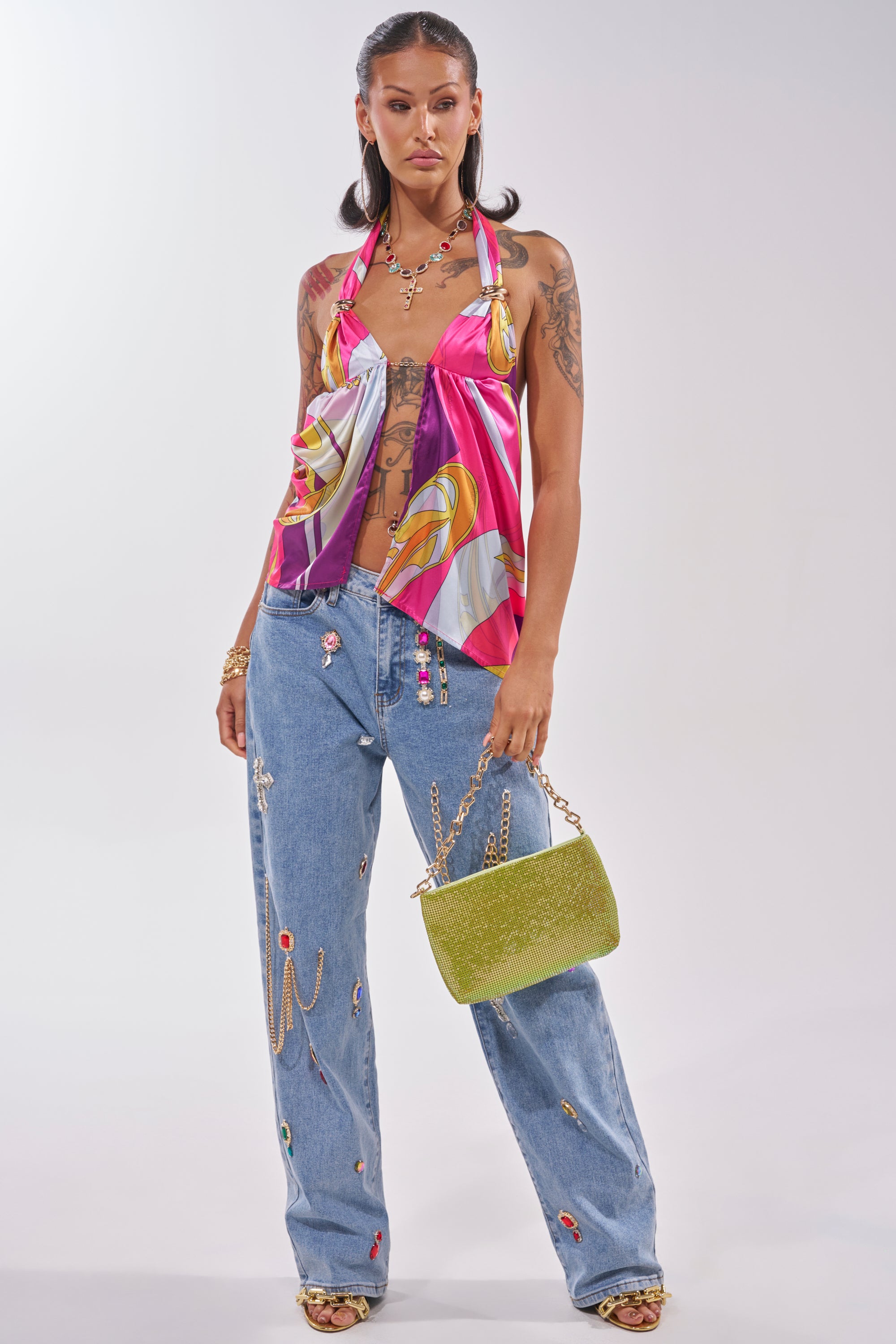 A woman poses in a satin halter top, FEELING THE DIFFERENCE EMBELLISHED RELAXED DENIM, gold heels, and a green textured bag. She wears gold jewelry, shows tattoos, and has her hair in loose waves. 6