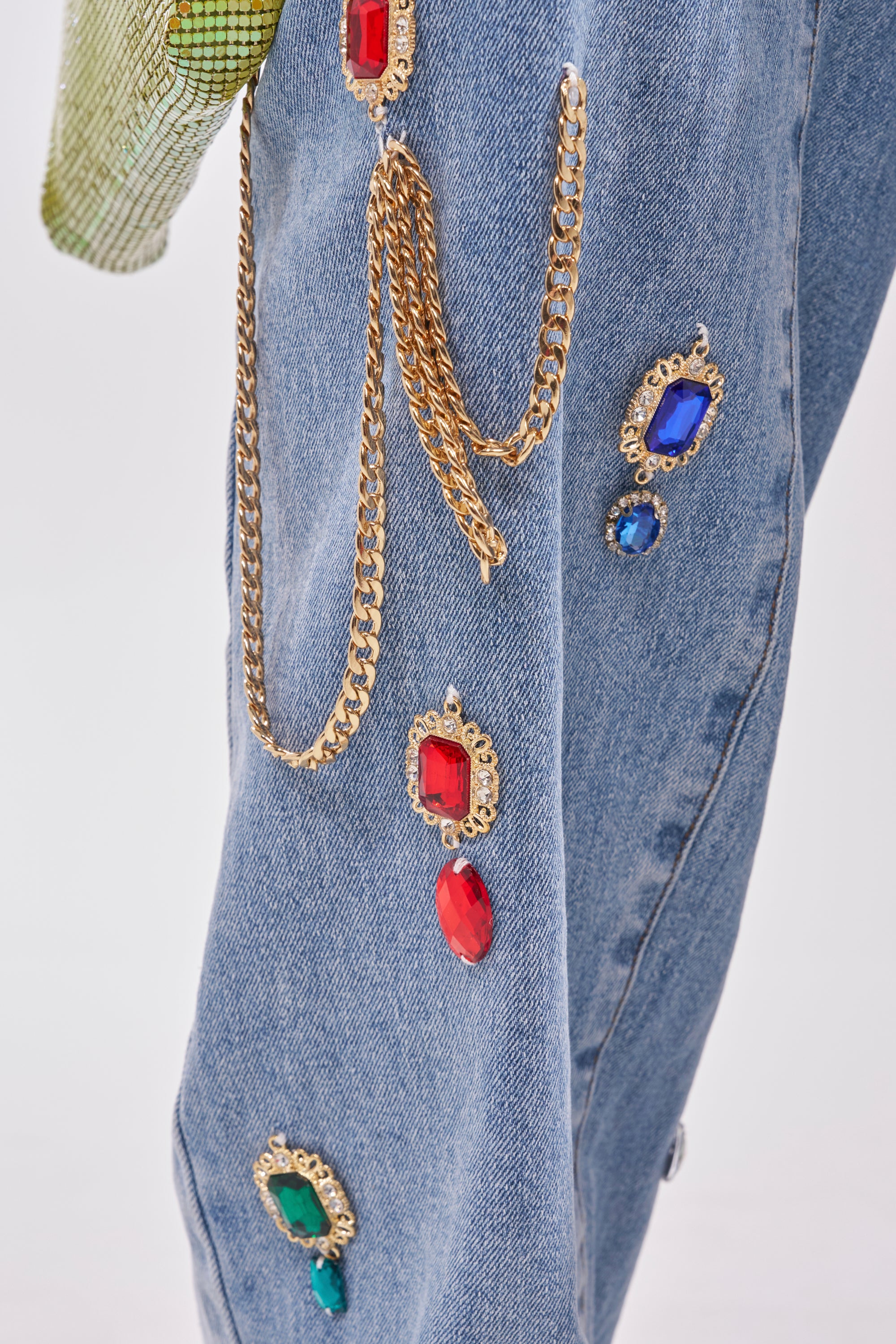 Close-up of FEELING THE DIFFERENCE EMBELLISHED RELAXED DENIM: blue jeans adorned with gold chains and colorful jeweled accents in ornate gold settings, plus a hint of green fabric at the top left. 8