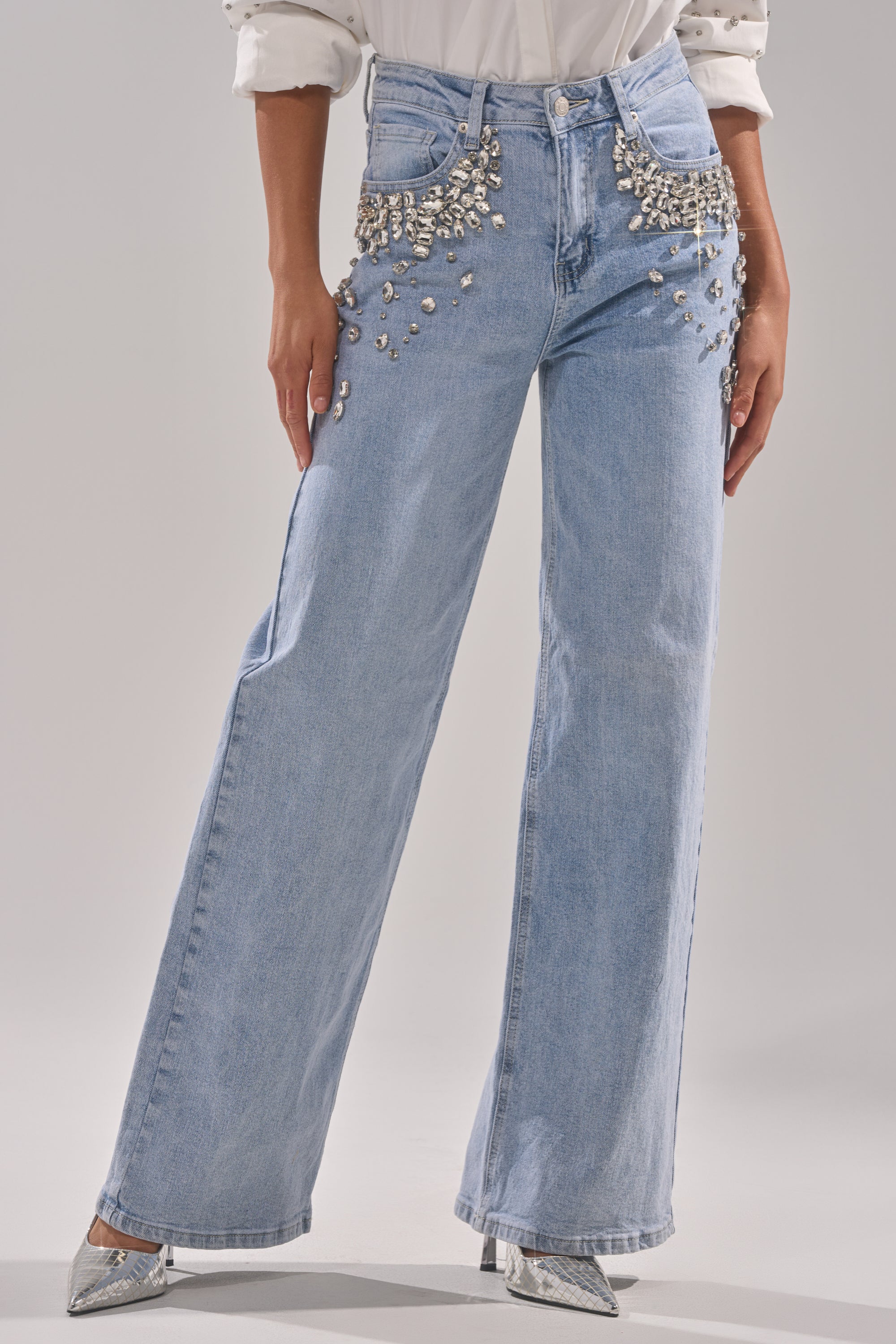 A person wearing the ITS THAT TIME EMBELLISHED RELAXED DENIM—light blue wide-leg jeans with silver embellishments on the hips and thighs—styled with a white shirt and silver pointed-toe heels.