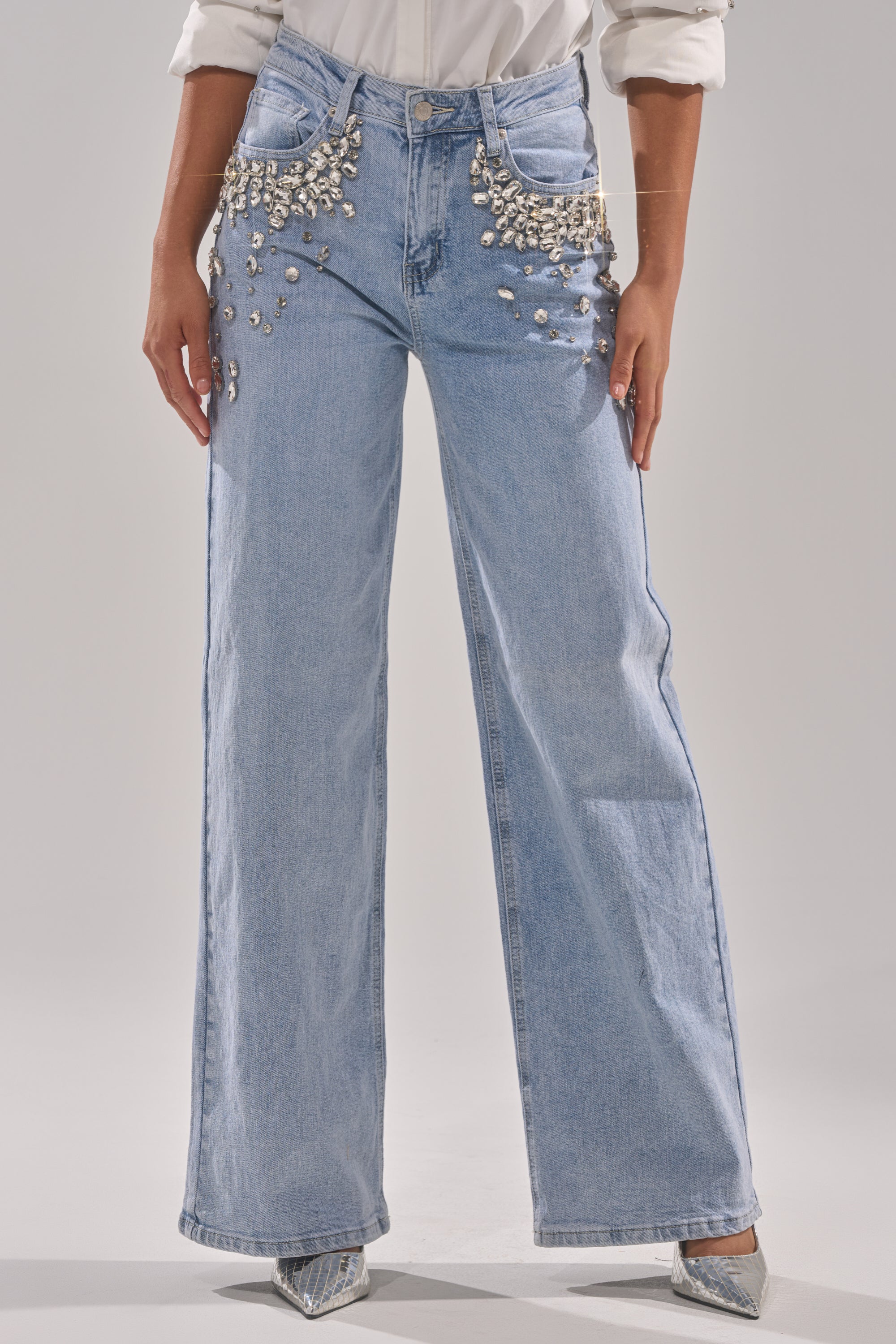 Someone wears the ITS THAT TIME EMBELLISHED RELAXED DENIM in light blue with silver rhinestones on the front pockets, styled with a tucked-in white shirt and pointed silver shoes. The upper body is not visible.