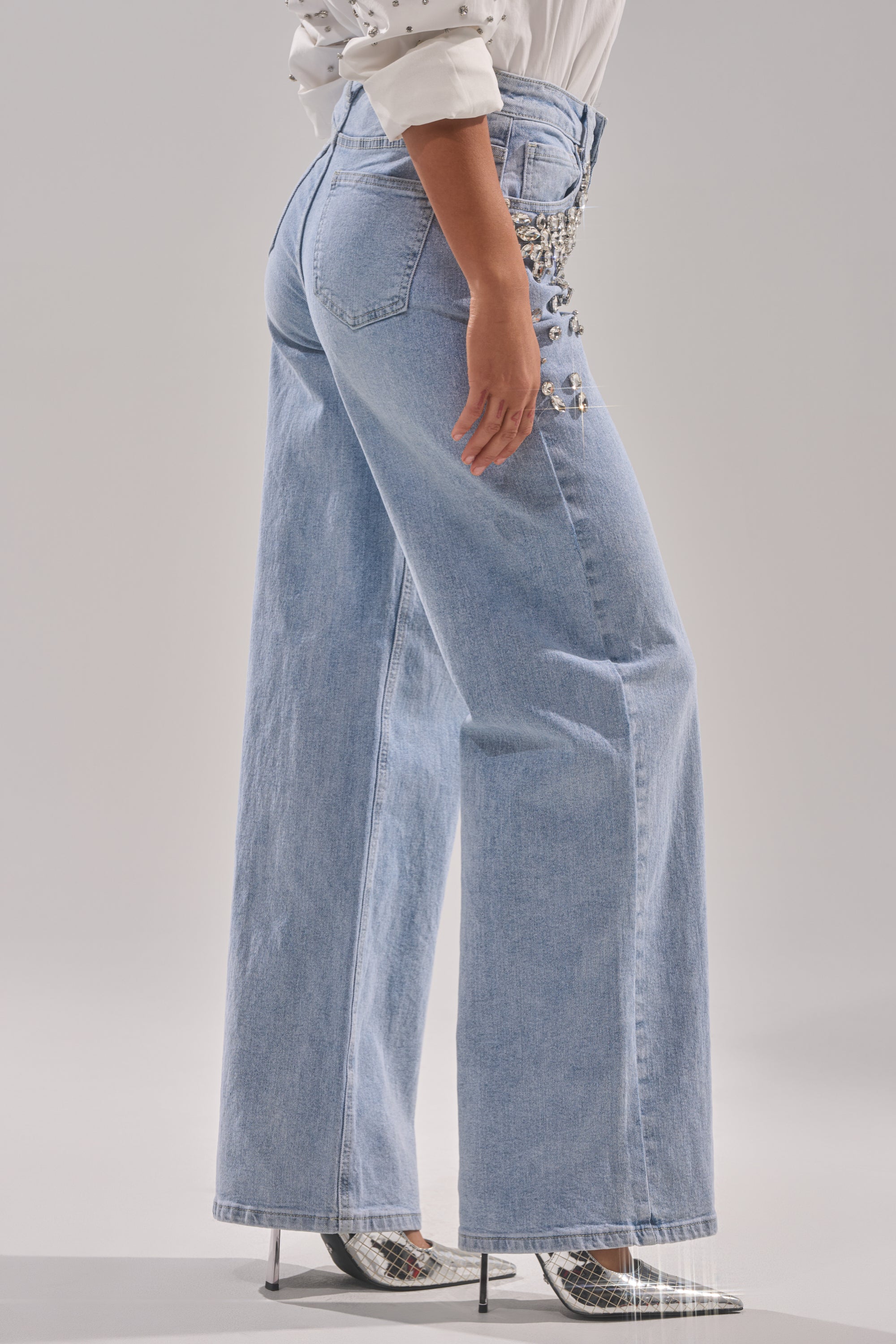 Someone wears the ITS THAT TIME EMBELLISHED RELAXED DENIM jeans, featuring high-rise wide legs and rhinestones on the side, with a white blouse and silver heels. The backdrop is plain and light-colored.