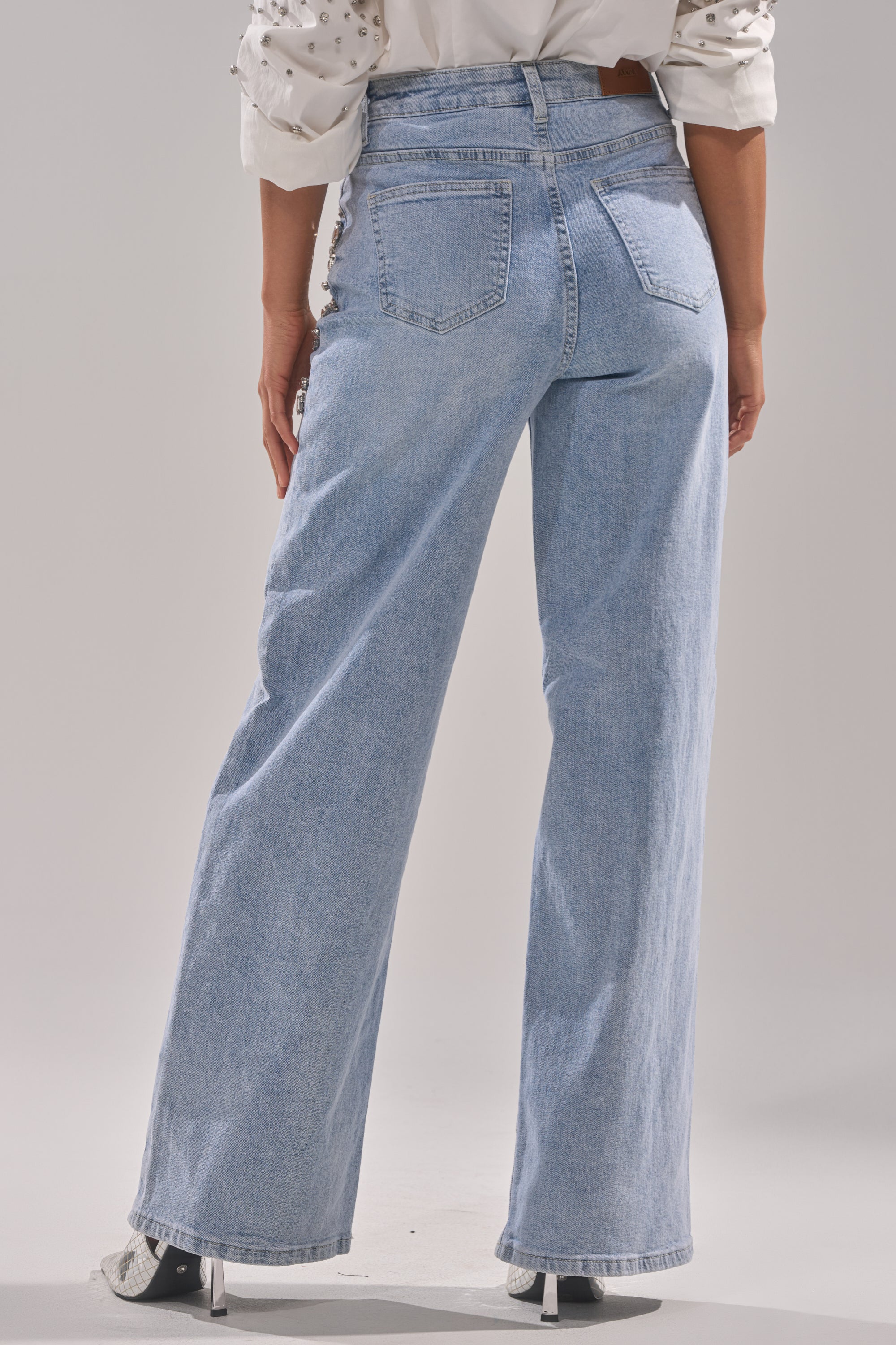 Wearing ITS THAT TIME EMBELLISHED RELAXED DENIM, a person in light blue high-waisted, wide-leg jeans and a white rolled-sleeve shirt stands with their back to the camera. The embellished denim lends subtle style to the relaxed outfit.