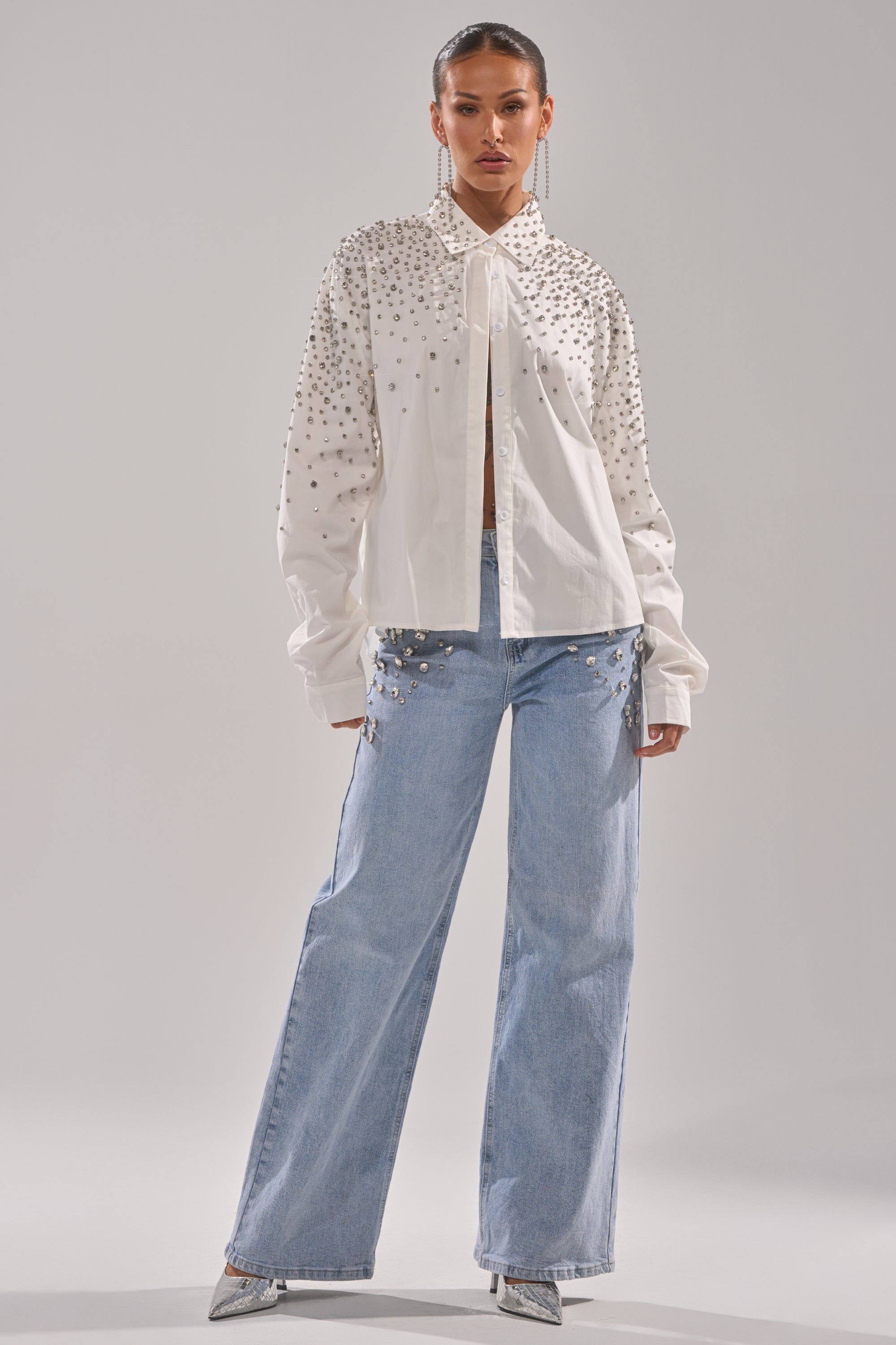 A woman faces forward wearing a white studded button-up shirt, ITS THAT TIME EMBELLISHED RELAXED DENIM jeans featuring studs, silver pointed-toe heels, long earrings, and has her hair pulled back.