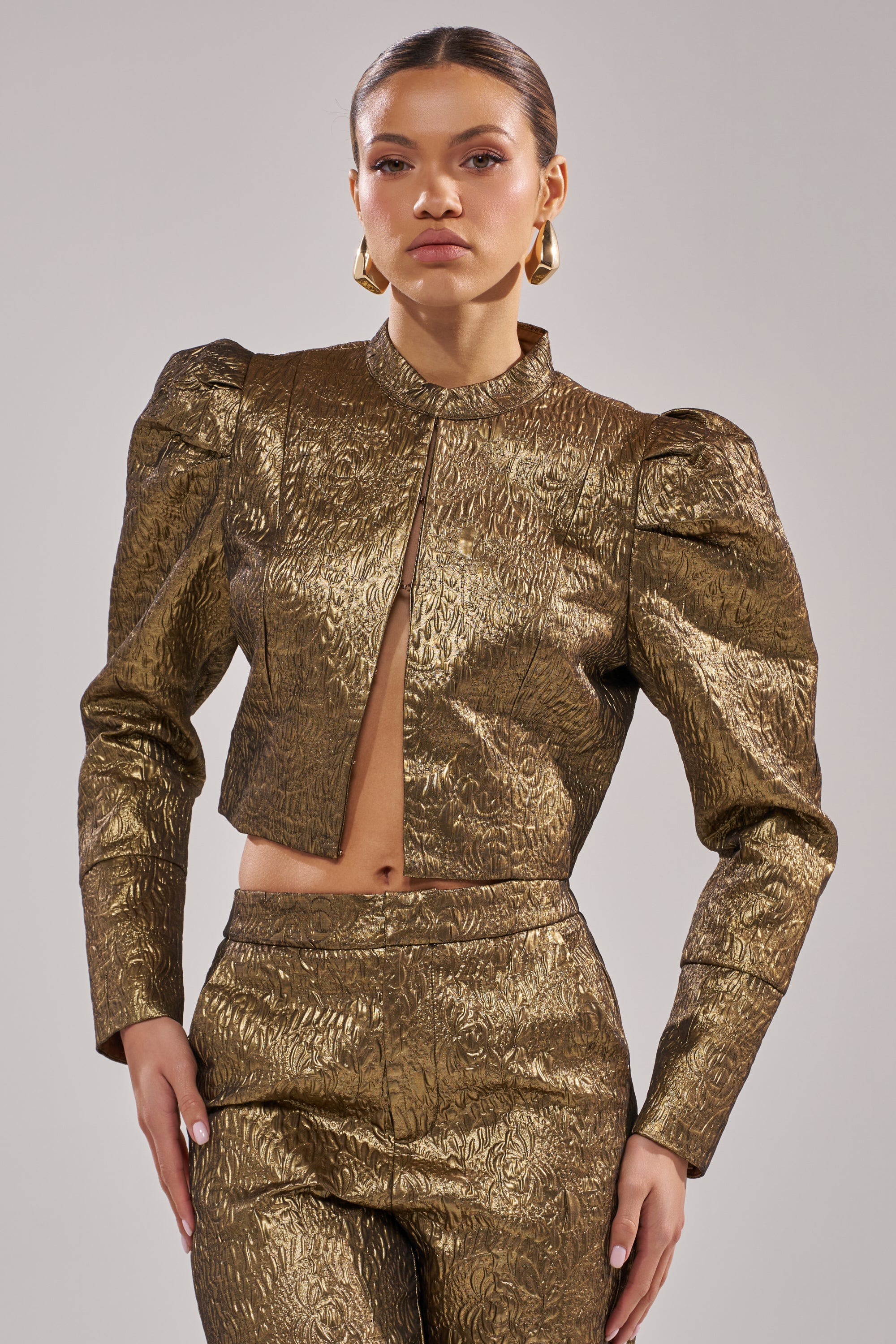 A woman poses confidently in the DAPHNE BROCADE BLAZER, featuring a shiny metallic gold finish, structured silhouette, and cropped cut. She pairs it with statement earrings and has her hair pulled back against a plain background.