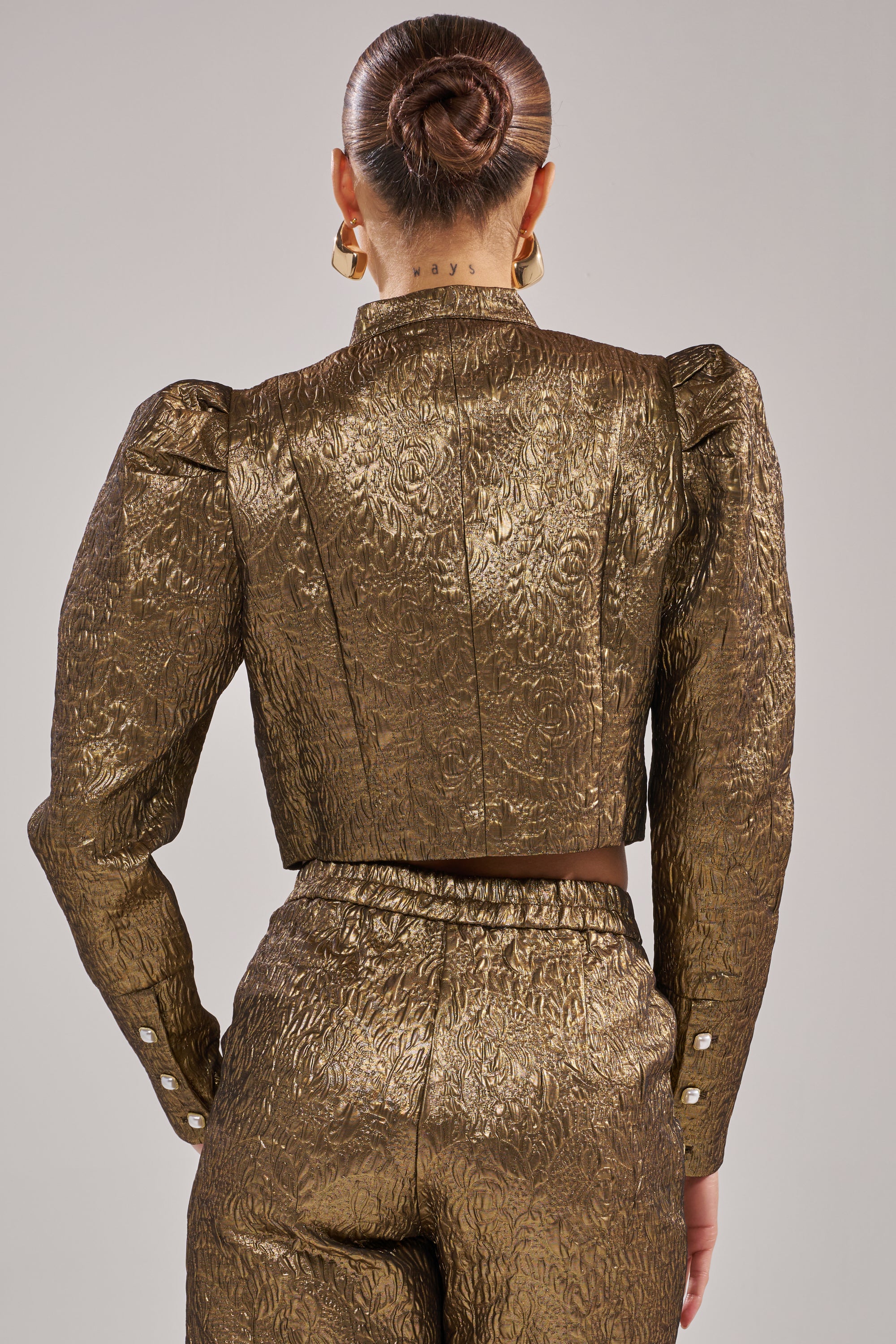 A sleek-bunned individual is seen from behind in the DAPHNE BROCADE BLAZER, a metallic gold piece with a structured silhouette, long sleeves, cuff buttons, and bold puffed shoulders, paired with matching pants.