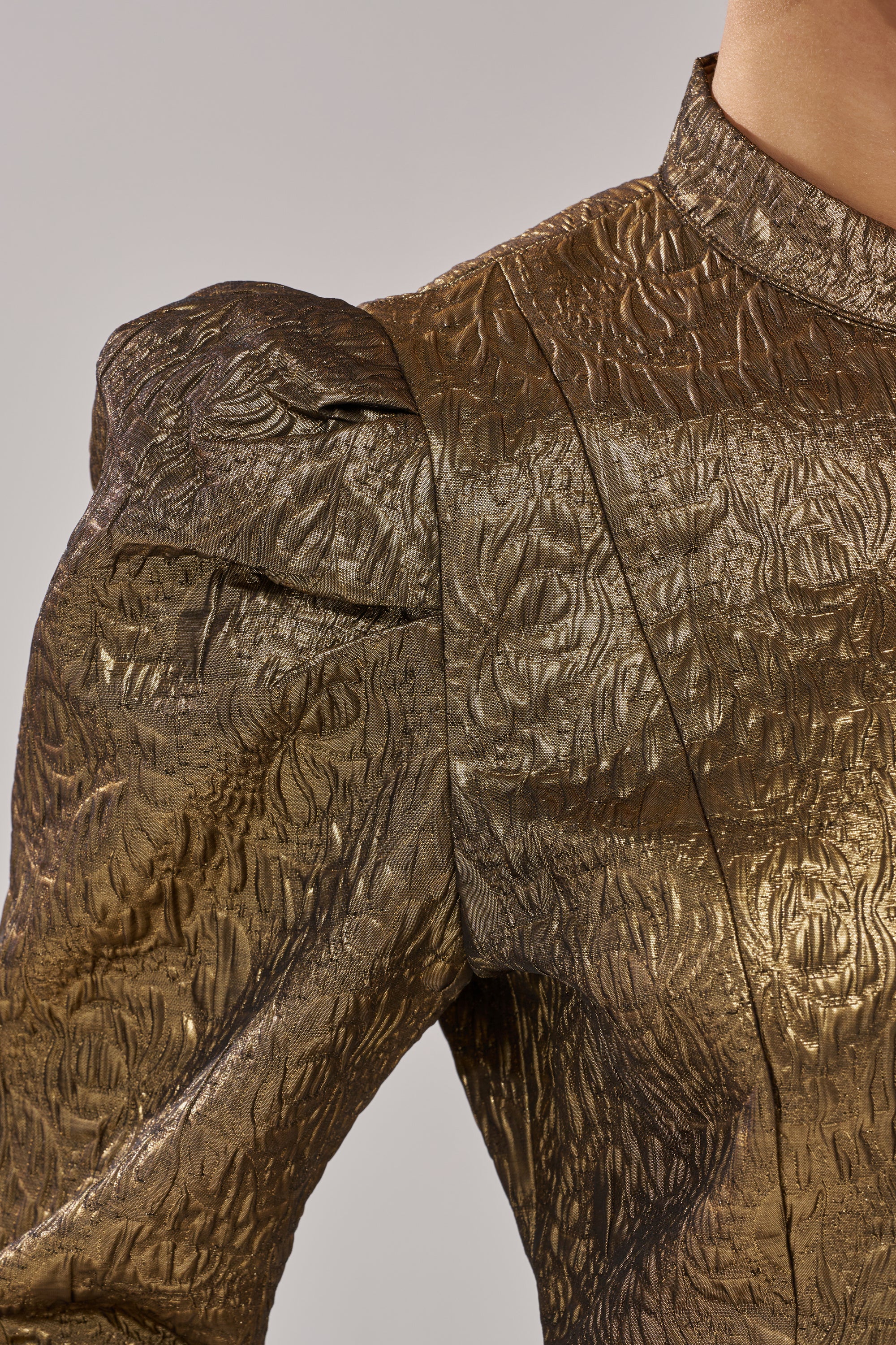 Close-up of a person wearing the DAPHNE BROCADE BLAZER in metallic gold with structured shoulders and intricate patterns, set against a plain light background. Only the upper torso and neck are visible.