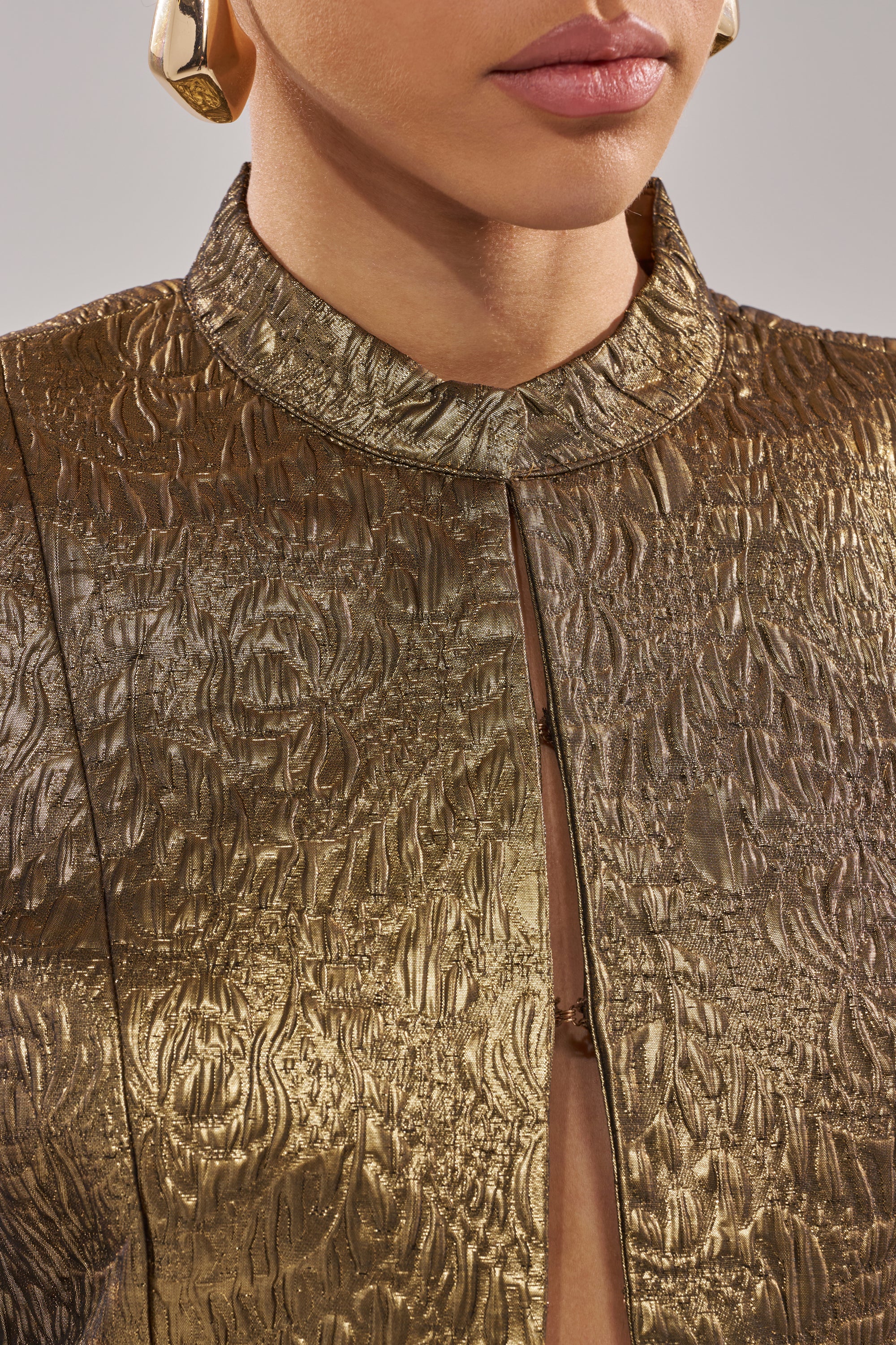 A close-up of a person wearing the DAPHNE BROCADE BLAZER in metallic gold with a high collar and front slit, styled with large gold hoop earrings. Only their lips and part of their face are visible.