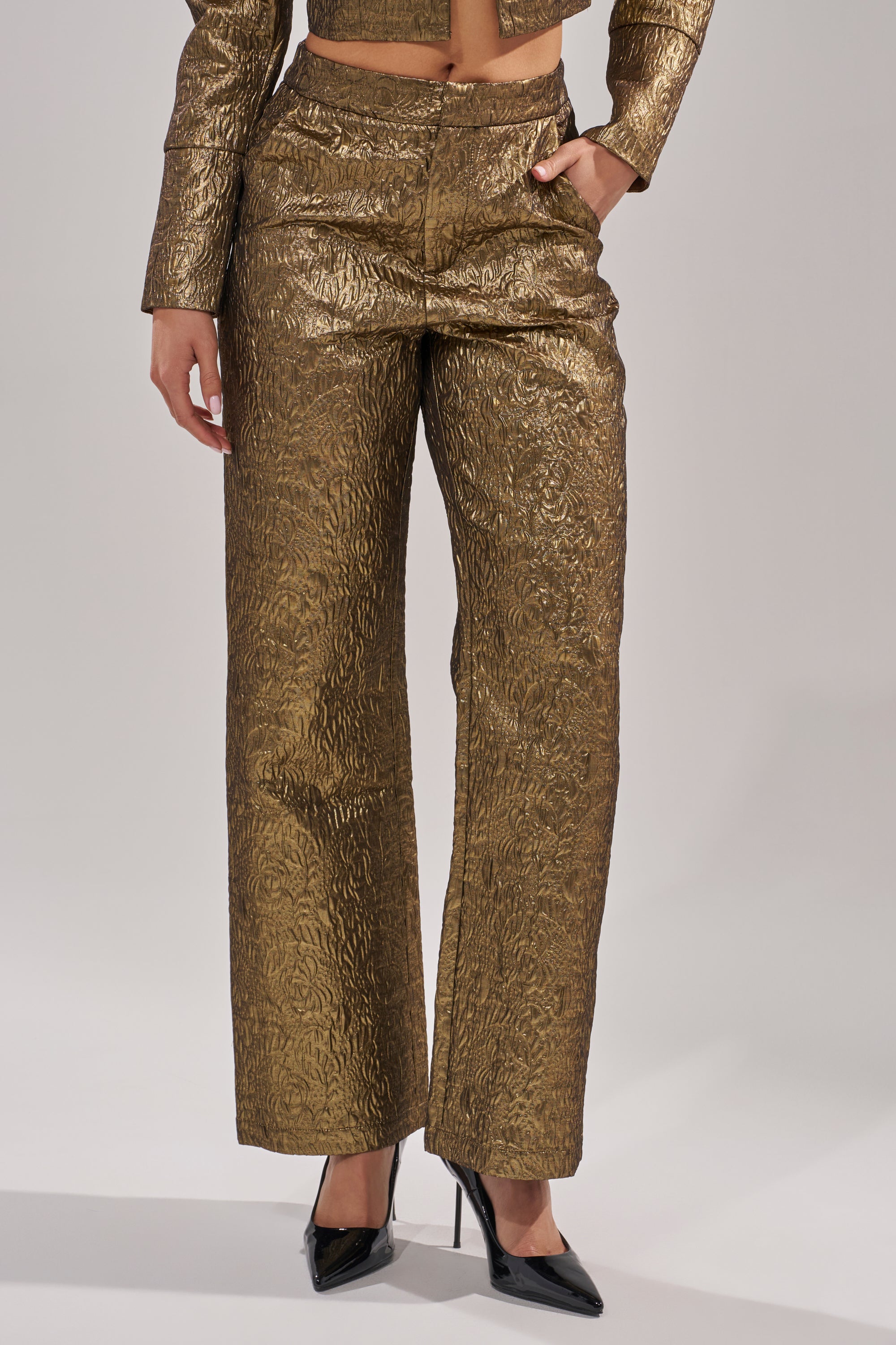 Wearing the DAPHNE BROCADE TROUSER and a matching jacket, with one hand in their pocket and pointed black high heels, a person stands against a plain light background. 0