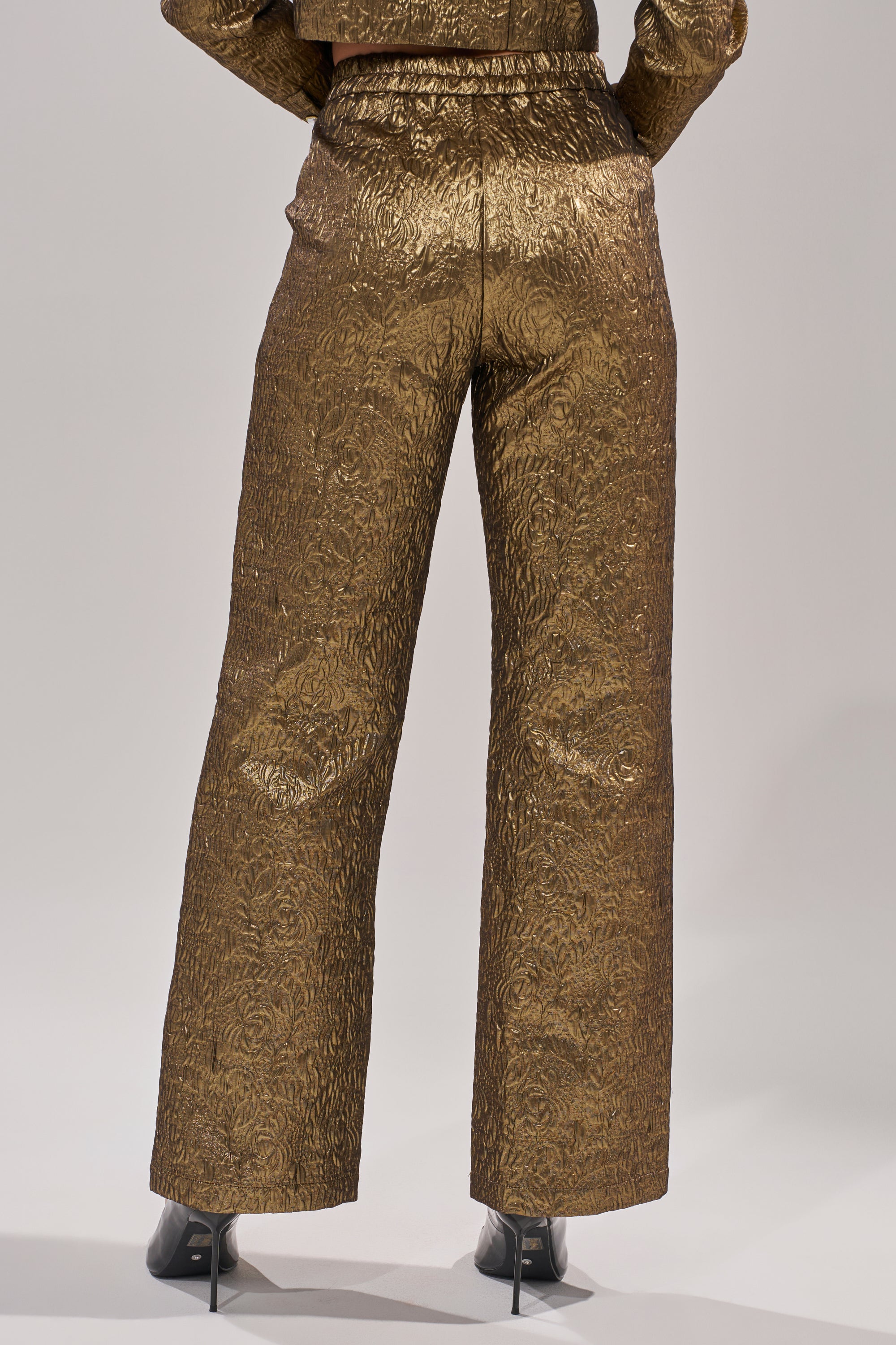 A person stands facing away, wearing the DAPHNE BROCADE TROUSER—high-waisted gold floral brocade pants with slightly flared legs—paired with black heeled shoes. 2
