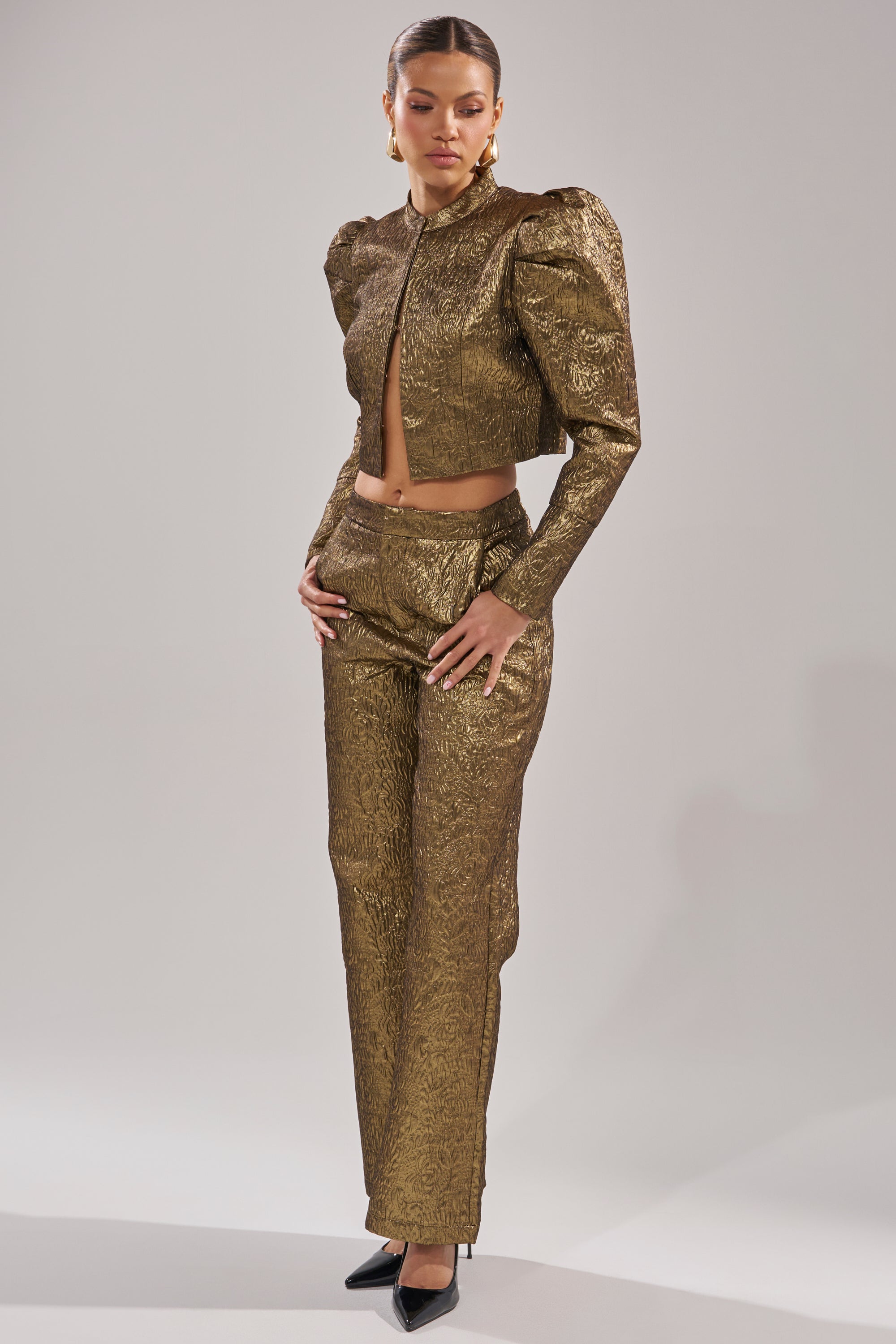 A woman poses confidently in DAPHNE BROCADE TROUSER, a textured, metallic gold high-waisted trouser paired with a cropped jacket. She stands with hands on her hips in pointed black heels against a plain gray background. 4