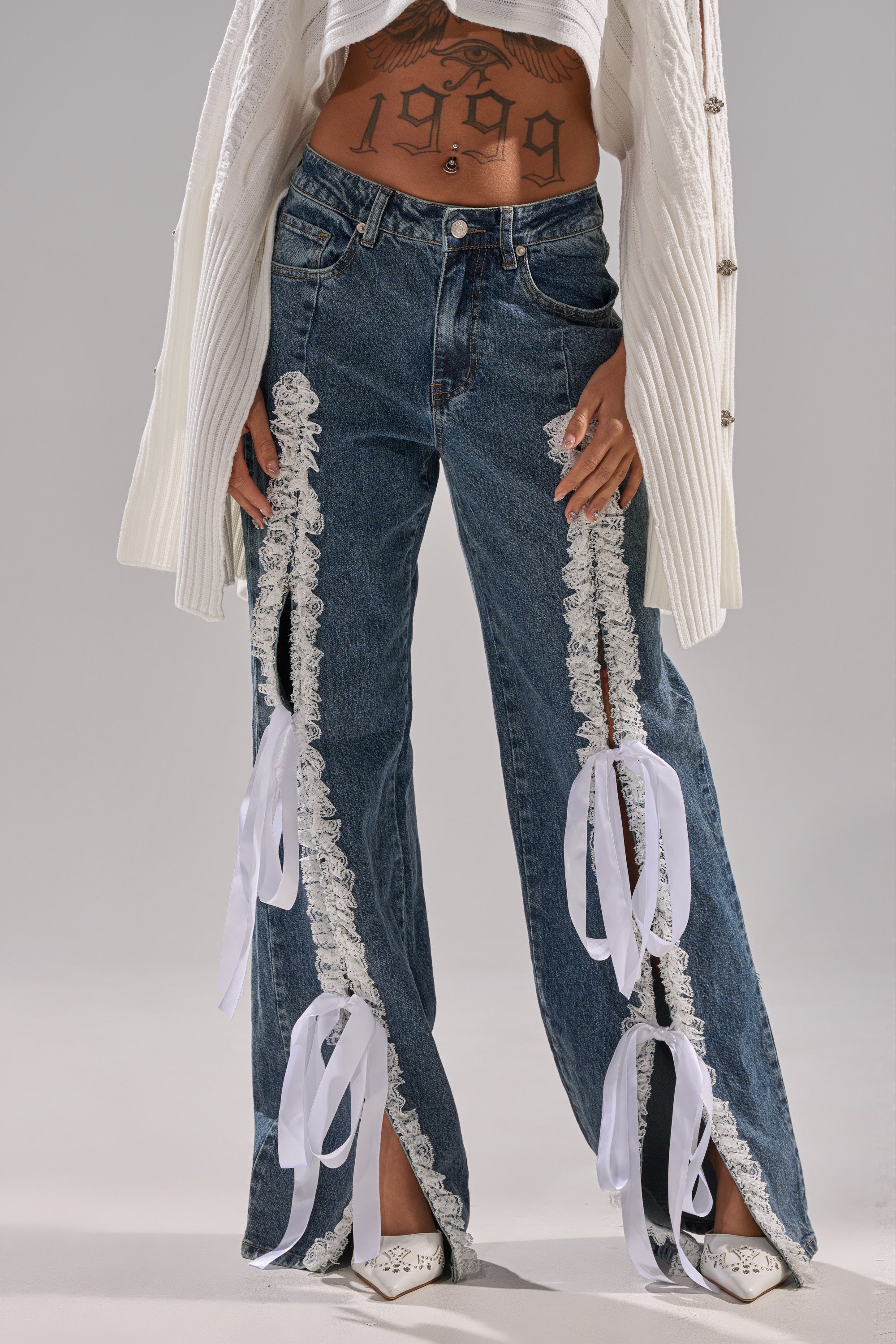 Someone wears TIE ME UP RELAXED DENIM jeans with lace trim, a white knit cardigan, white pointed-toe shoes, and a visible "1999" tattoo. The blue-denim look is photographed from mid-torso to feet.