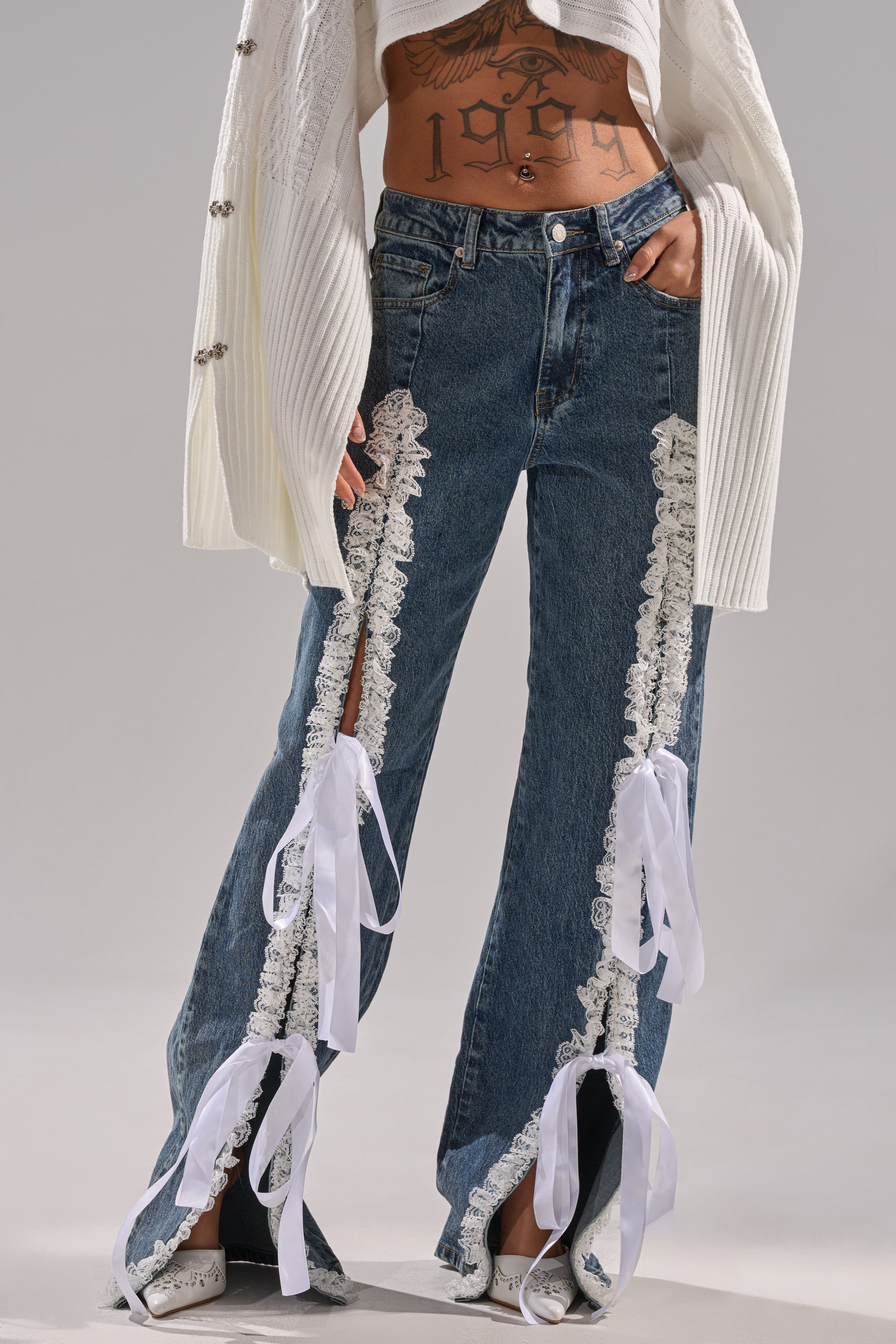 Someone wears the TIE ME UP RELAXED DENIM, featuring lace trim and white ribbon bows on front slits, styled with a cropped white sweater and white shoes. Their midriff, showing a "1999" tattoo, is visible.