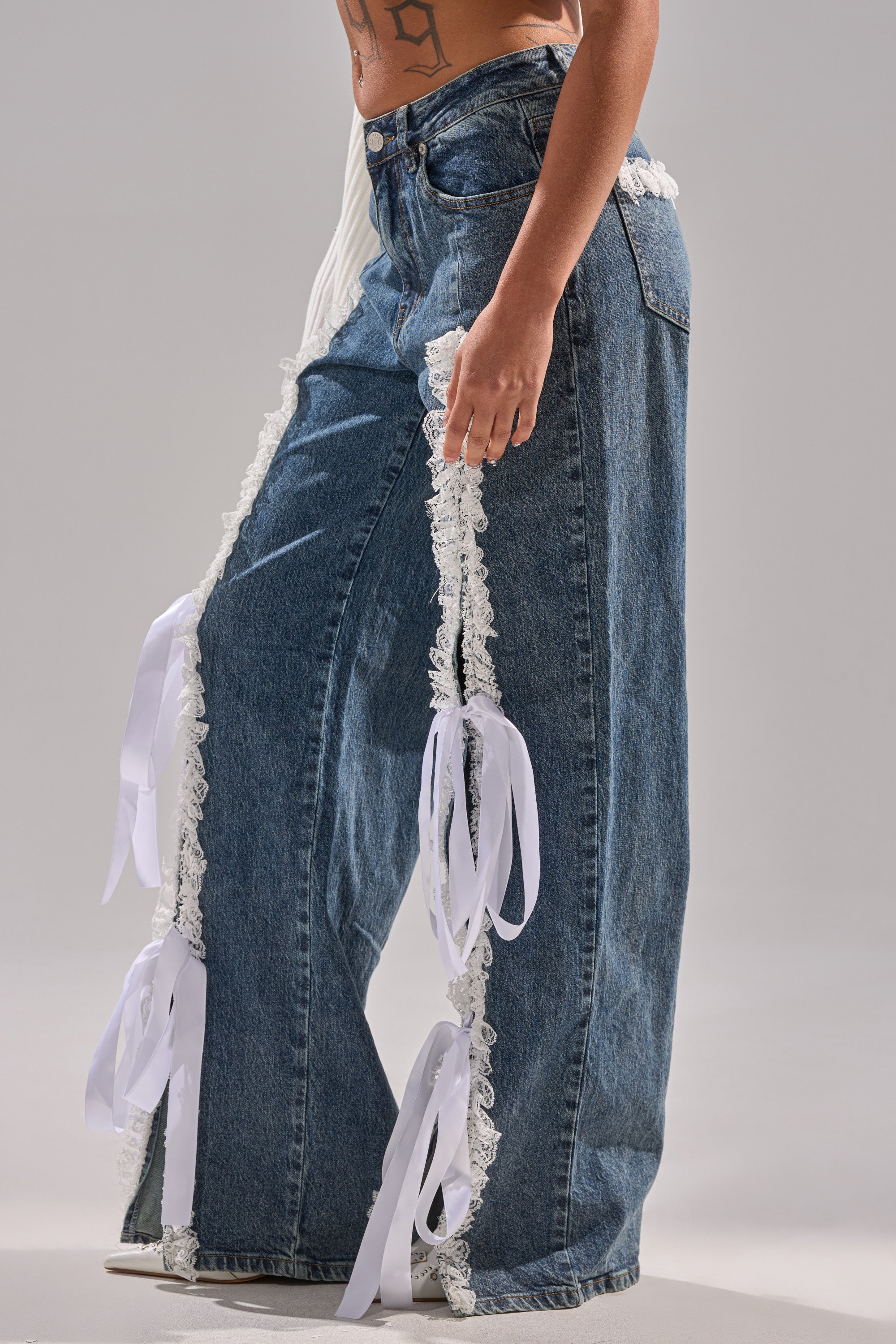 Wearing the TIE ME UP RELAXED DENIM, featuring high-rise lace-trimmed jeans with white ribbon bows on the sides, a model stands against a plain gray background, highlighting the relaxed and stylish fit visible from the lower torso and legs.