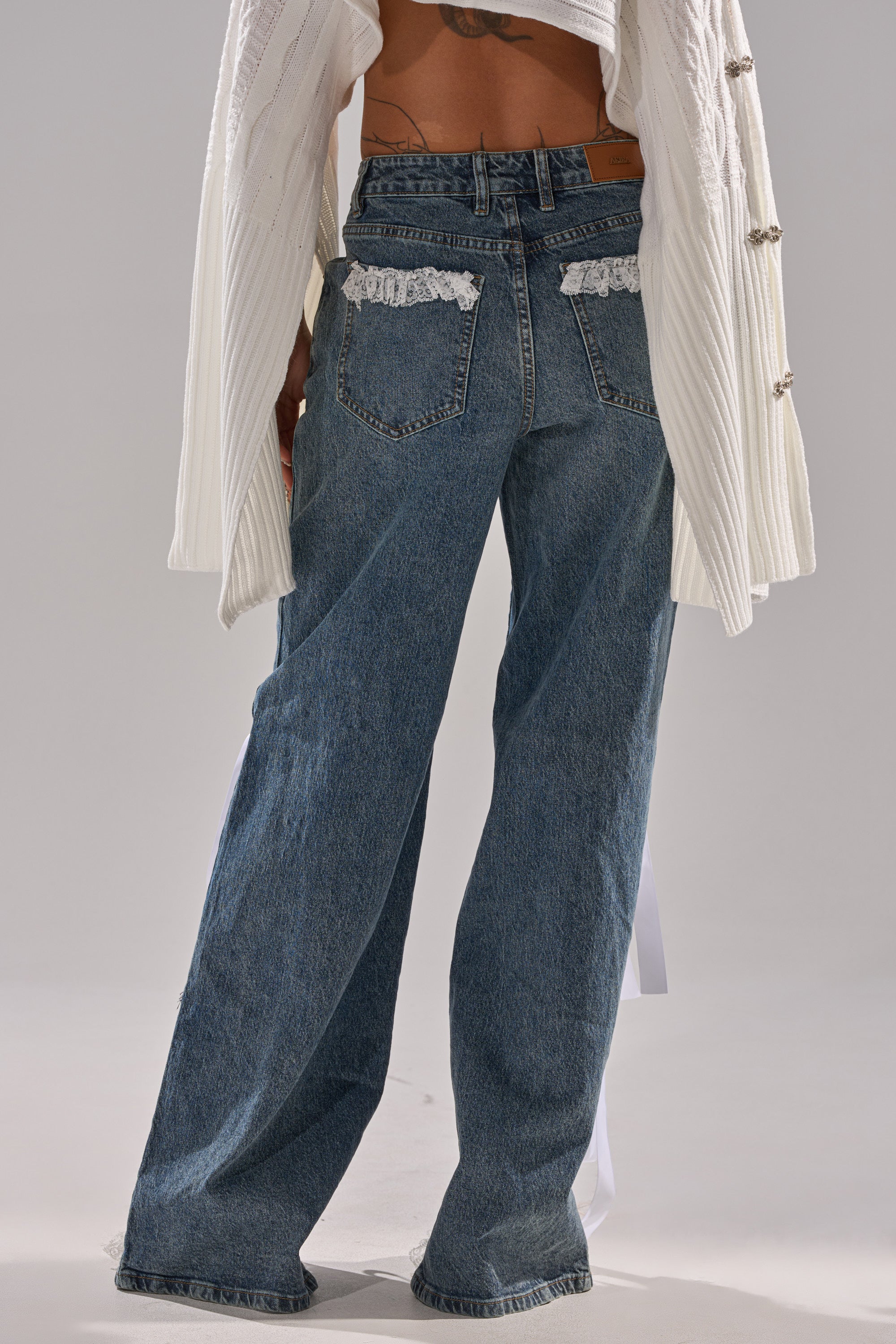 A person wears the TIE ME UP RELAXED DENIM—lace-trimmed, high-rise relaxed jeans—paired with a white textured long-sleeve top. The photo captures the back view, emphasizing the lower back and legs.