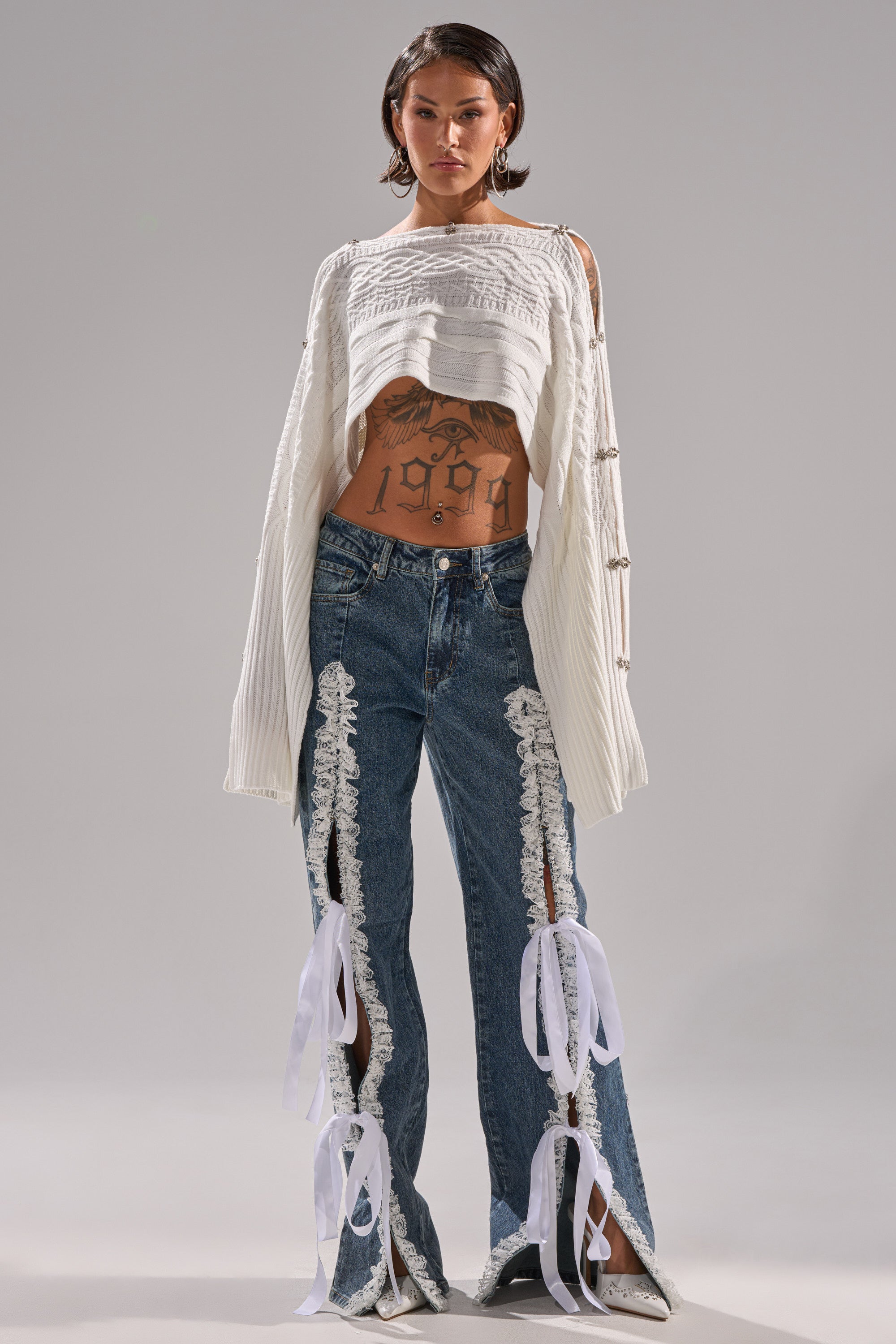 A woman stands against a plain background wearing TIE ME UP RELAXED DENIM—high-rise jeans with lace trim and white bows—paired with a cropped white sweater, white shoes, hoop earrings, and short hair, revealing her midriff tattoos.