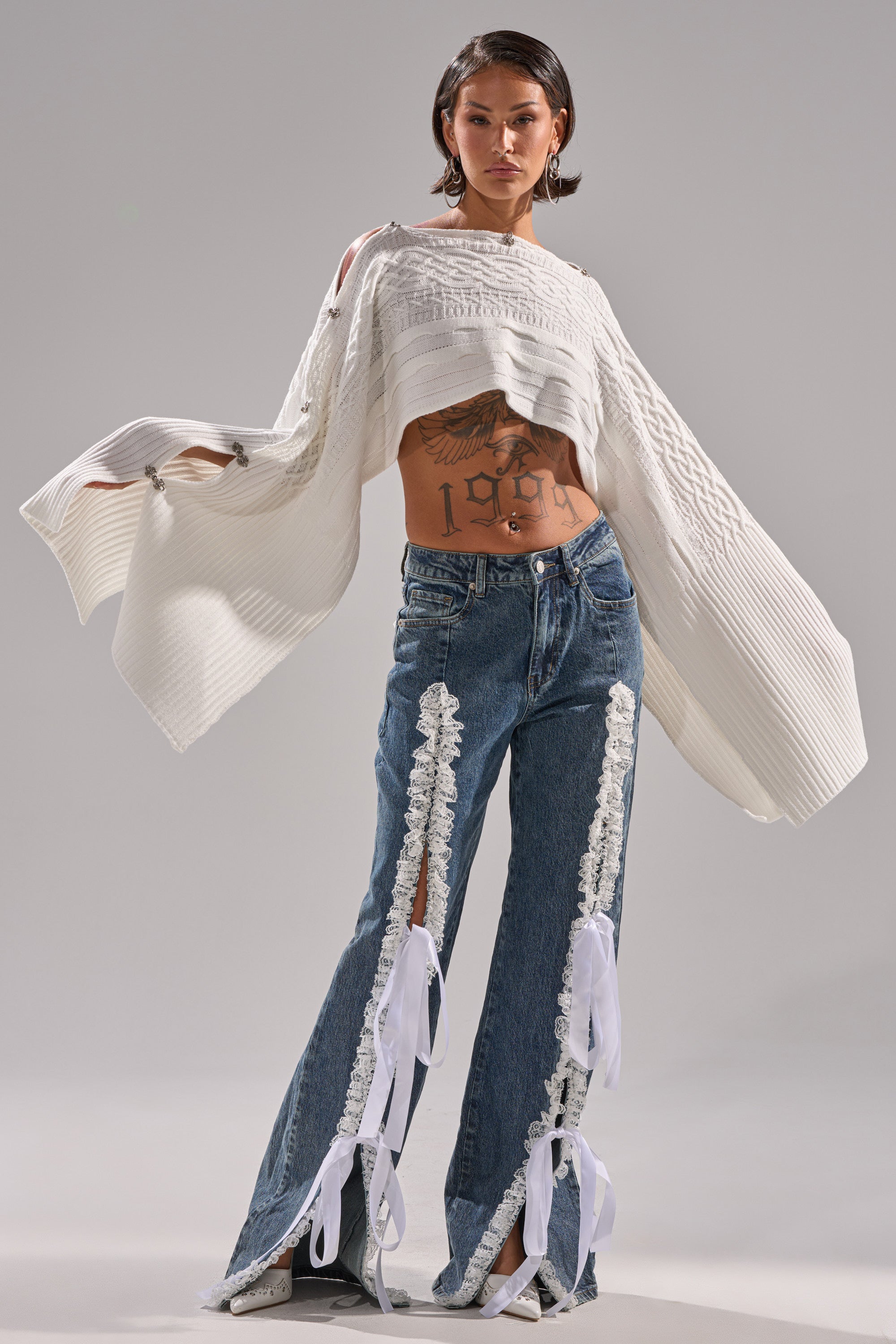 A woman models the "TIE ME UP RELAXED DENIM," featuring lace-trimmed jeans with ribbon details along the front seams and hem slits, paired with a white textured poncho-style top with wide sleeves, revealing her tattooed midriff.