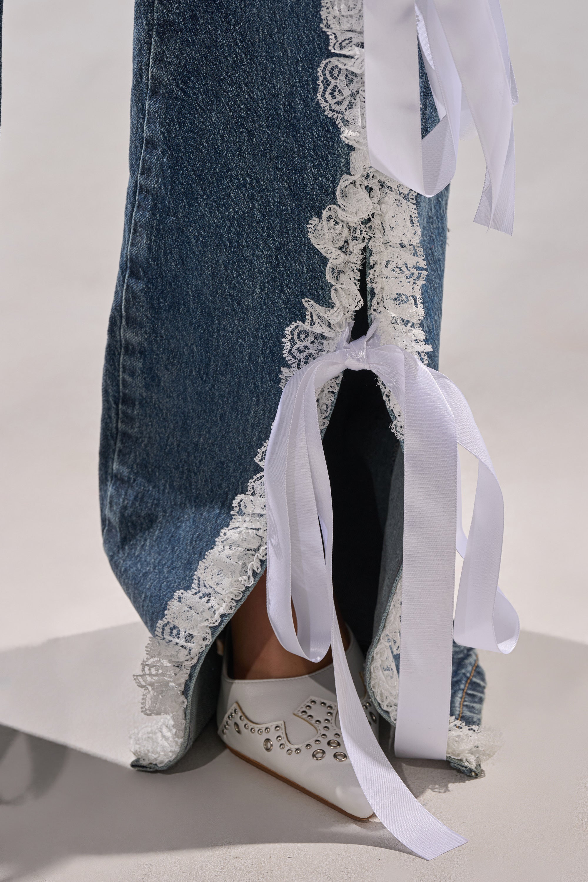 A close-up of the TIE ME UP RELAXED DENIM, featuring lace trim and long white ribbon bows, styled with white pointed shoes embellished with metallic studs.