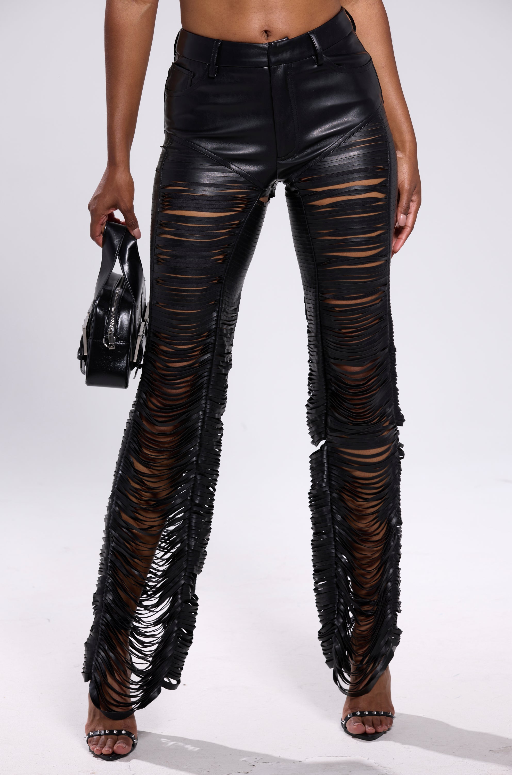 A person in SAME OL' MISTAKES DISTRESSED LEATHER PANT—high-rise skinny faux leather pants with horizontal slits—holding a small black handbag, wearing open-toe heels, against a plain white background. 1