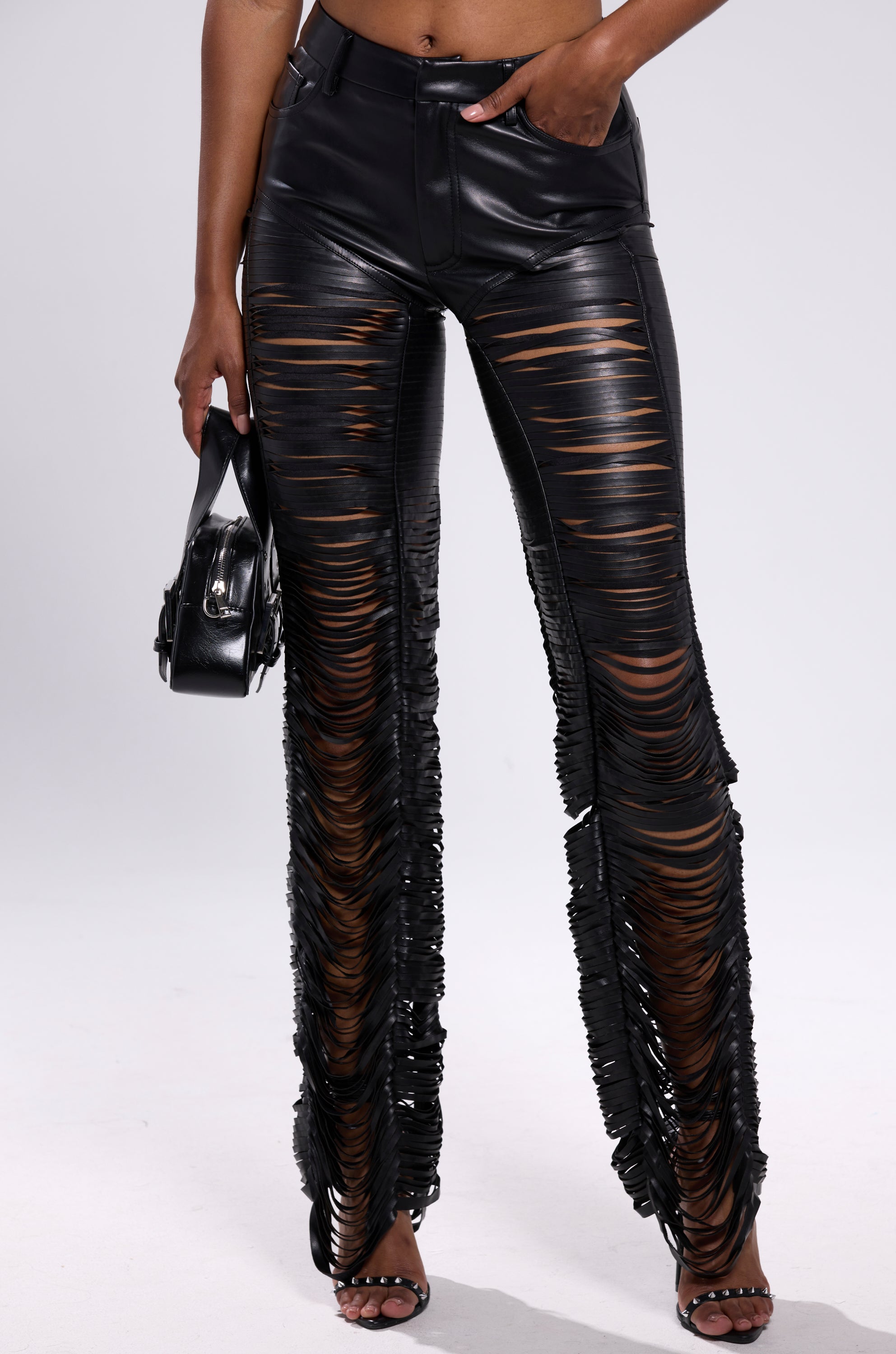 A person wearing SAME OL' MISTAKES DISTRESSED LEATHER PANT—high-rise, black faux leather with horizontal cut-out slits—paired with high-heeled sandals and a small black handbag, against a plain white background. 2