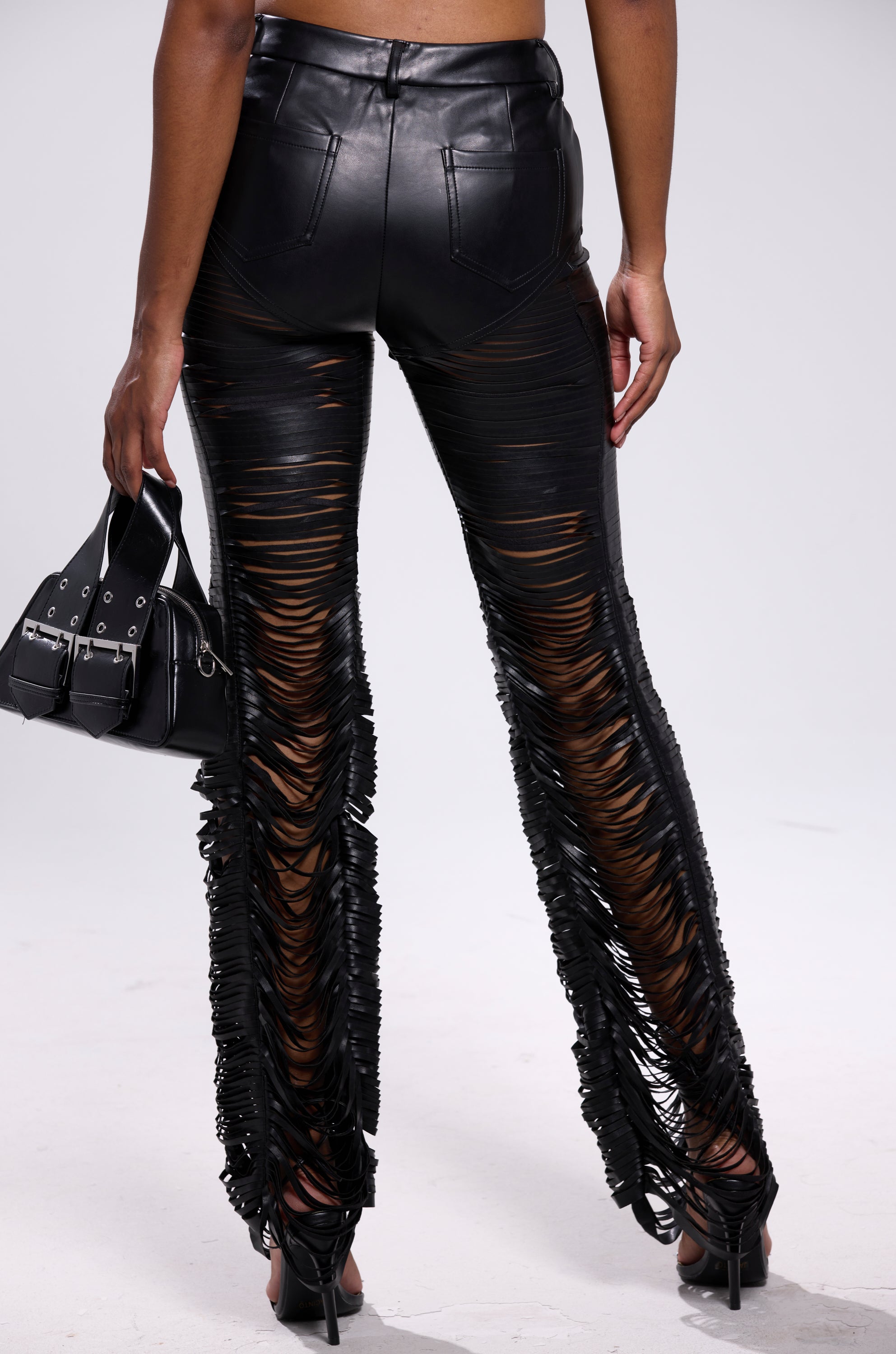Seen from behind, a person wears the SAME OL' MISTAKES DISTRESSED LEATHER PANT—black, high-rise skinny faux leather pants with bold horizontal slits—paired with high-heeled sandals and a small black handbag. 4