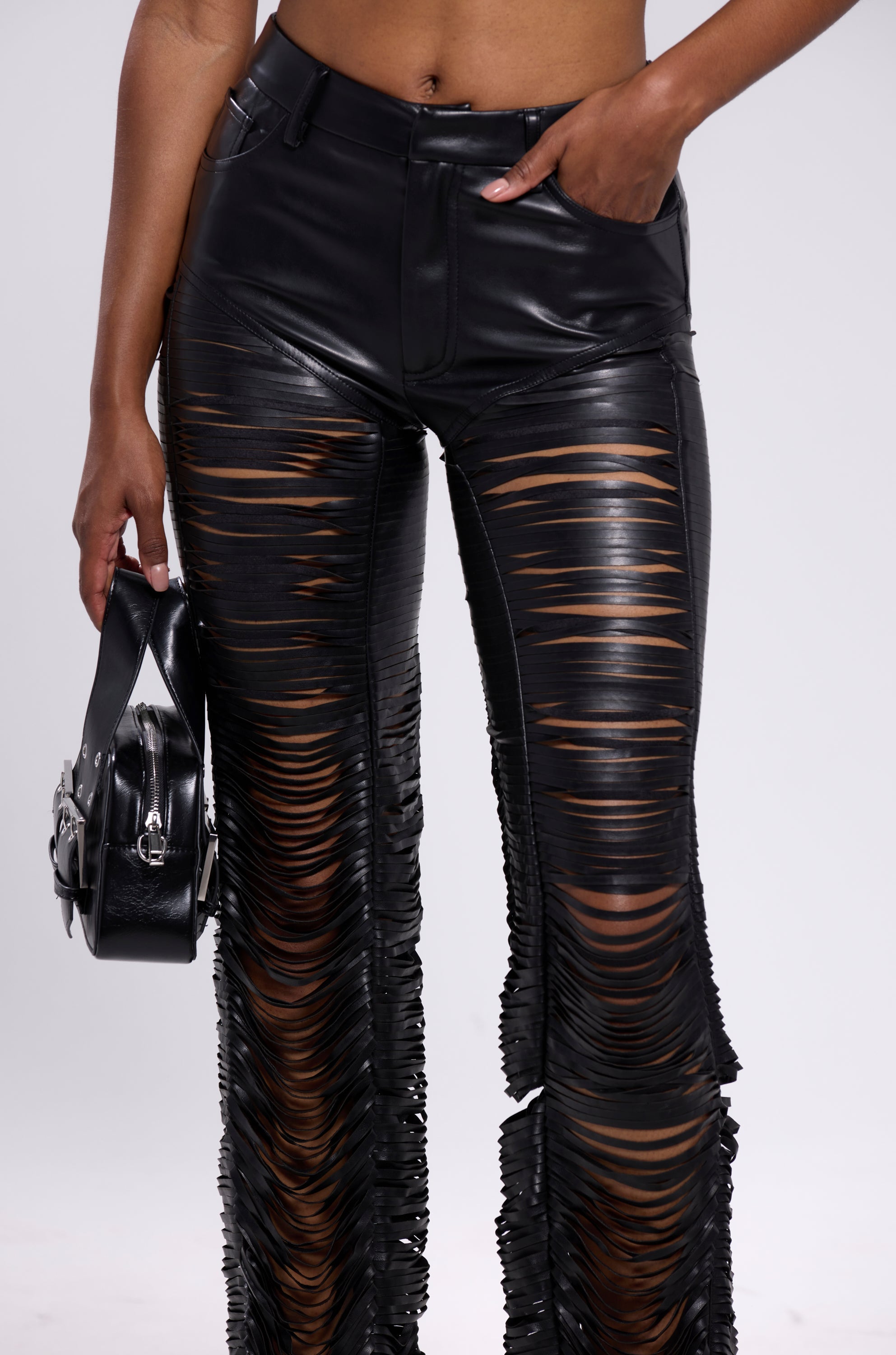 A person wearing the SAME OL' MISTAKES DISTRESSED LEATHER PANT—high-rise, black faux leather skinny pants with horizontal slashes—holds a black handbag and poses with one hand in their pocket against a plain background. 7