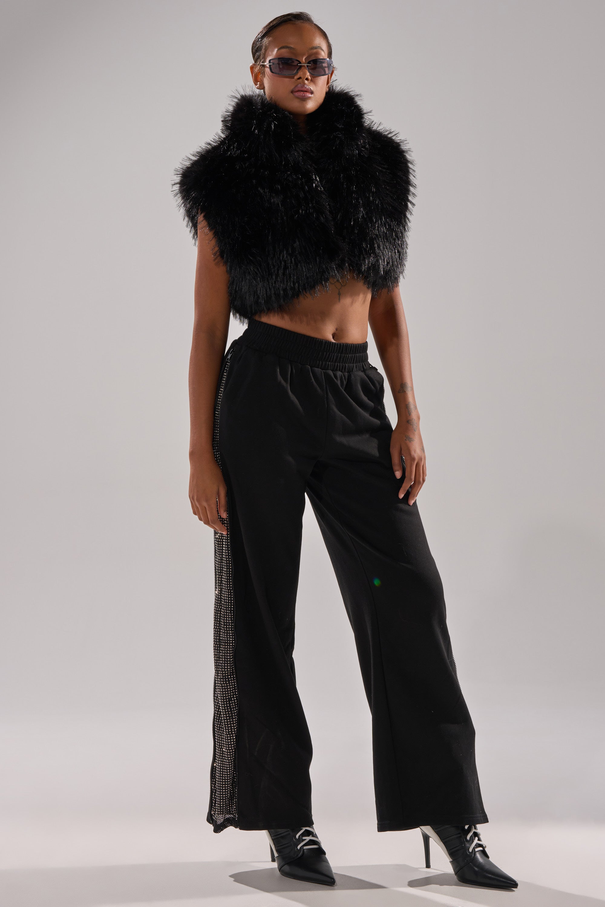 A person stands confidently in a black faux fur cropped vest, OPEN UP WIDE LEG JOGGER WITH RHINESTONE pants, black pointed-toe ankle boots, and sunglasses against a plain gray background. 0