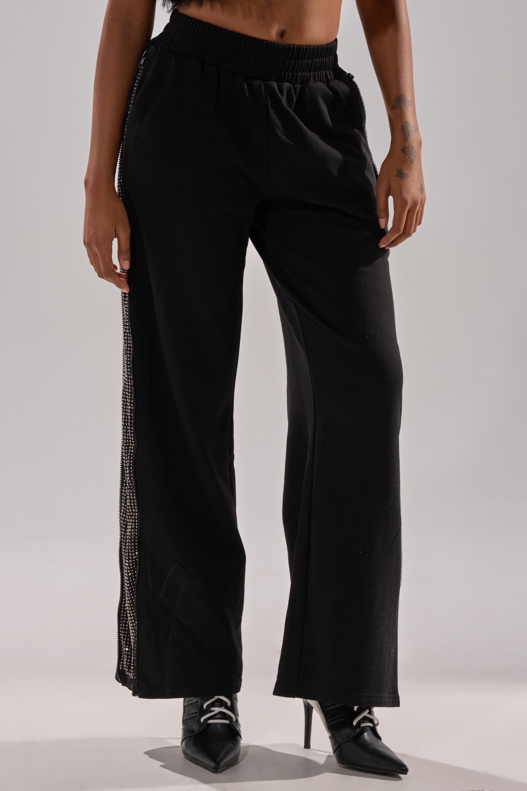 A person wears the OPEN UP WIDE LEG JOGGER WITH RHINESTONE—black wide-leg joggers with rhinestone side detailing—paired with black pointed-toe heeled shoes, standing against a plain light background. 3