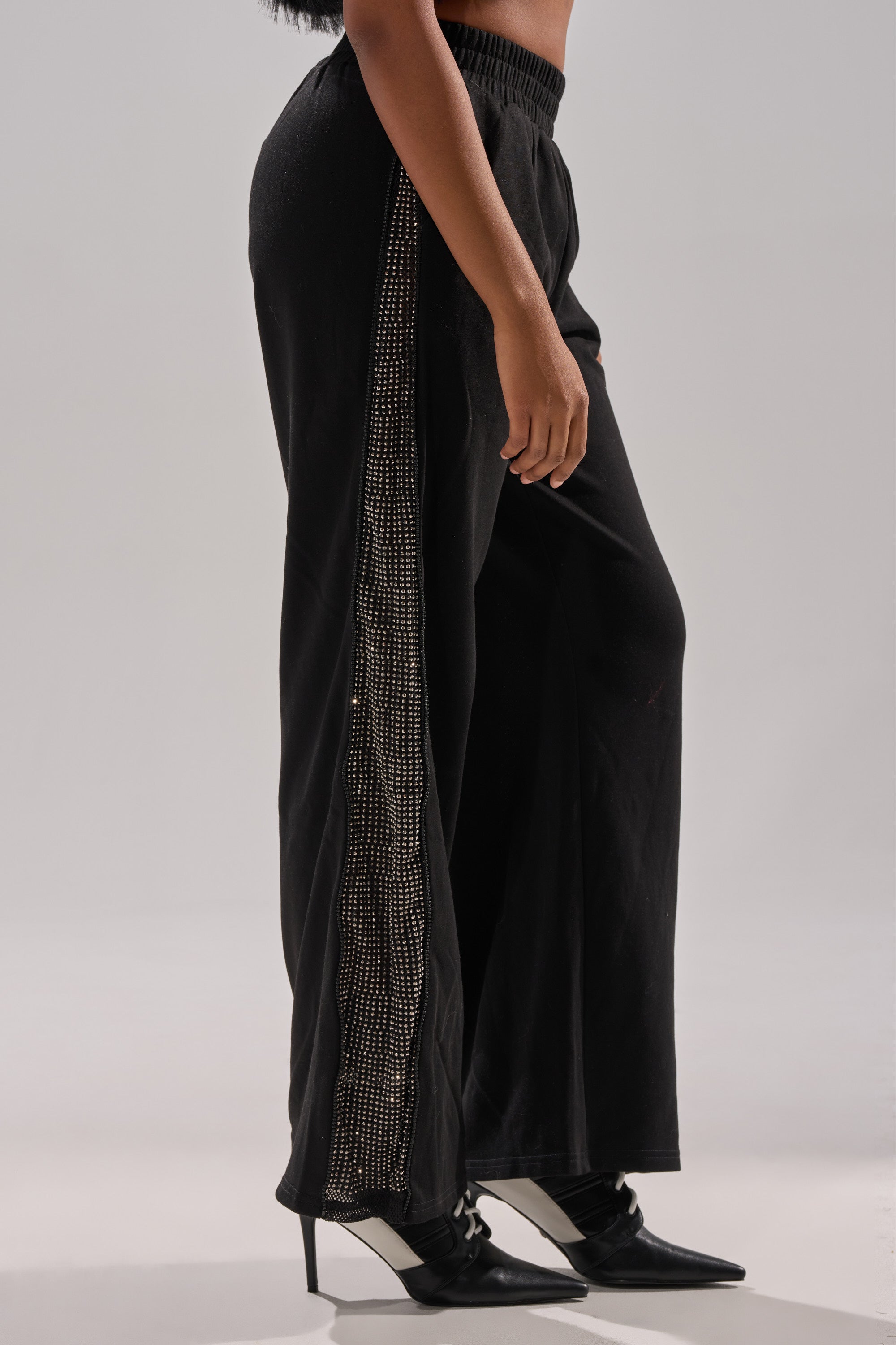 A person wears the OPEN UP WIDE LEG JOGGER WITH RHINESTONE featuring a sparkly side stripe, paired with black pointed-toe heels, standing against a plain background. 4
