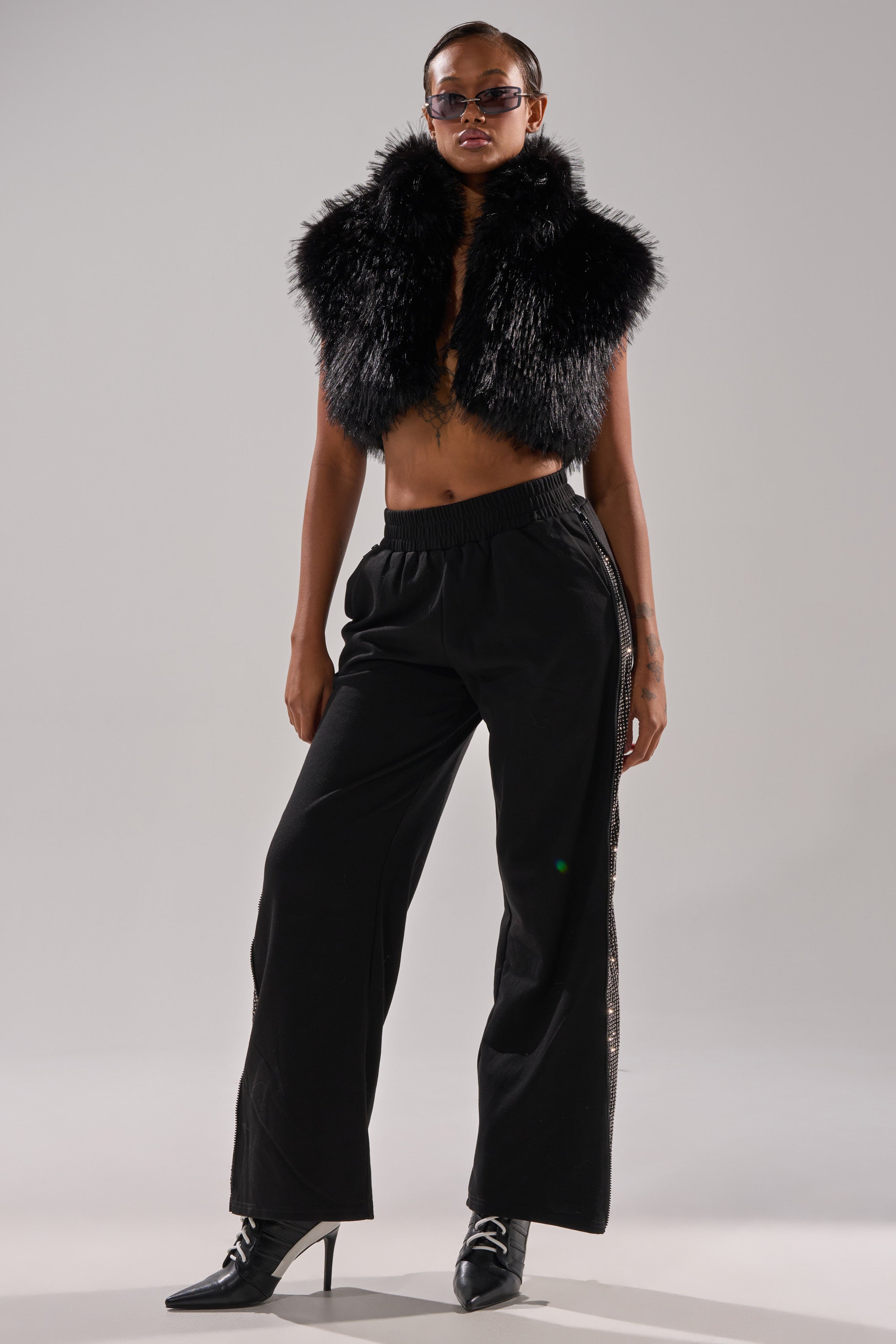A person poses confidently wearing a black fur vest, OPEN UP WIDE LEG JOGGER WITH RHINESTONE pants, pointed black boots, and sunglasses against a plain light gray background. 7