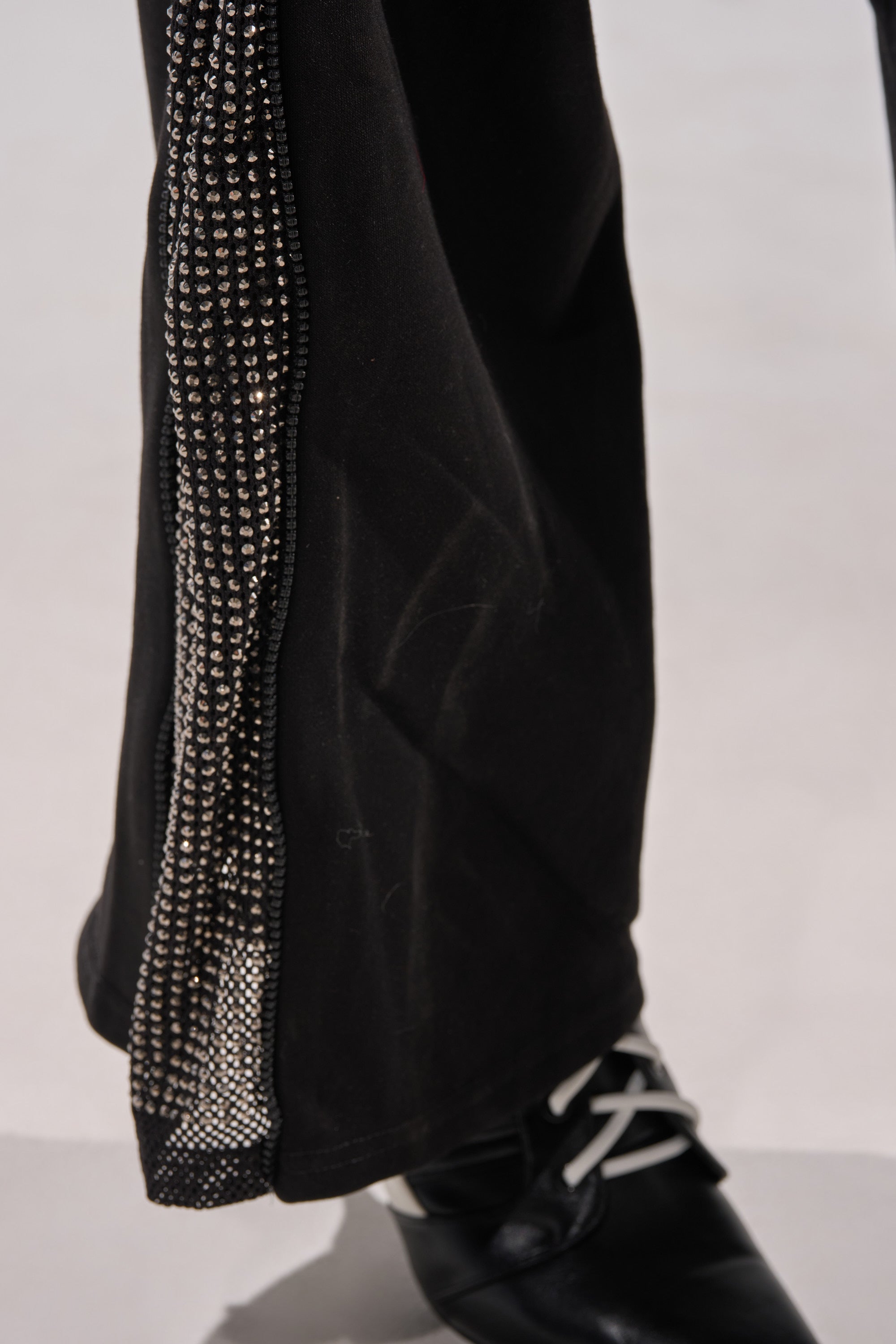 Close-up of the OPEN UP WIDE LEG JOGGER WITH RHINESTONE, showing a black pant leg with a vertical row of sparkling rhinestones down the side, paired with a black shoe with white laces, set against a light background. 8