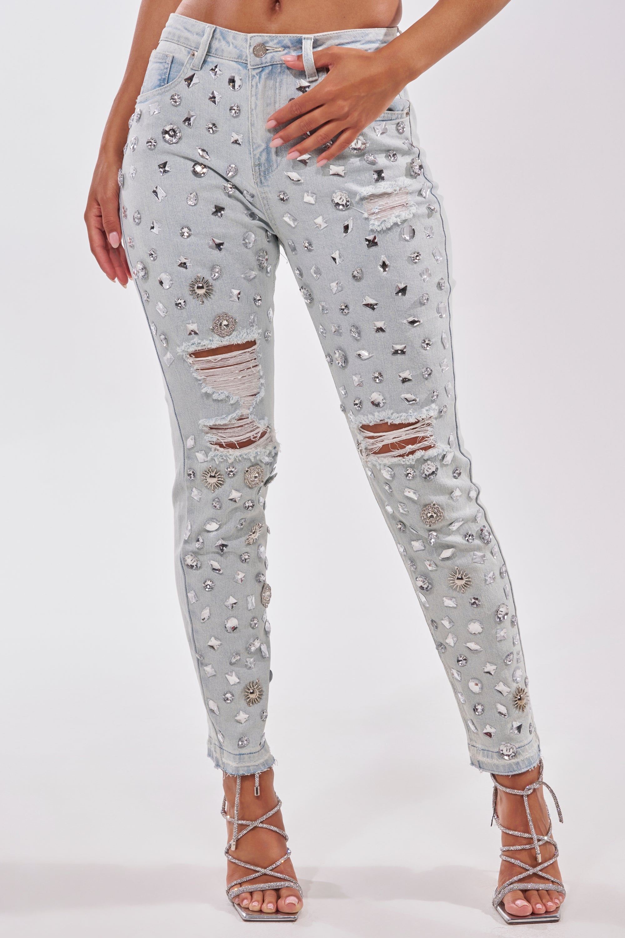 A person wearing DAMN GIRL EMBELLISHED SKINNY DENIM, light blue jeans with silver sequin details, paired with strappy high-heeled sandals, stands against a plain white background.