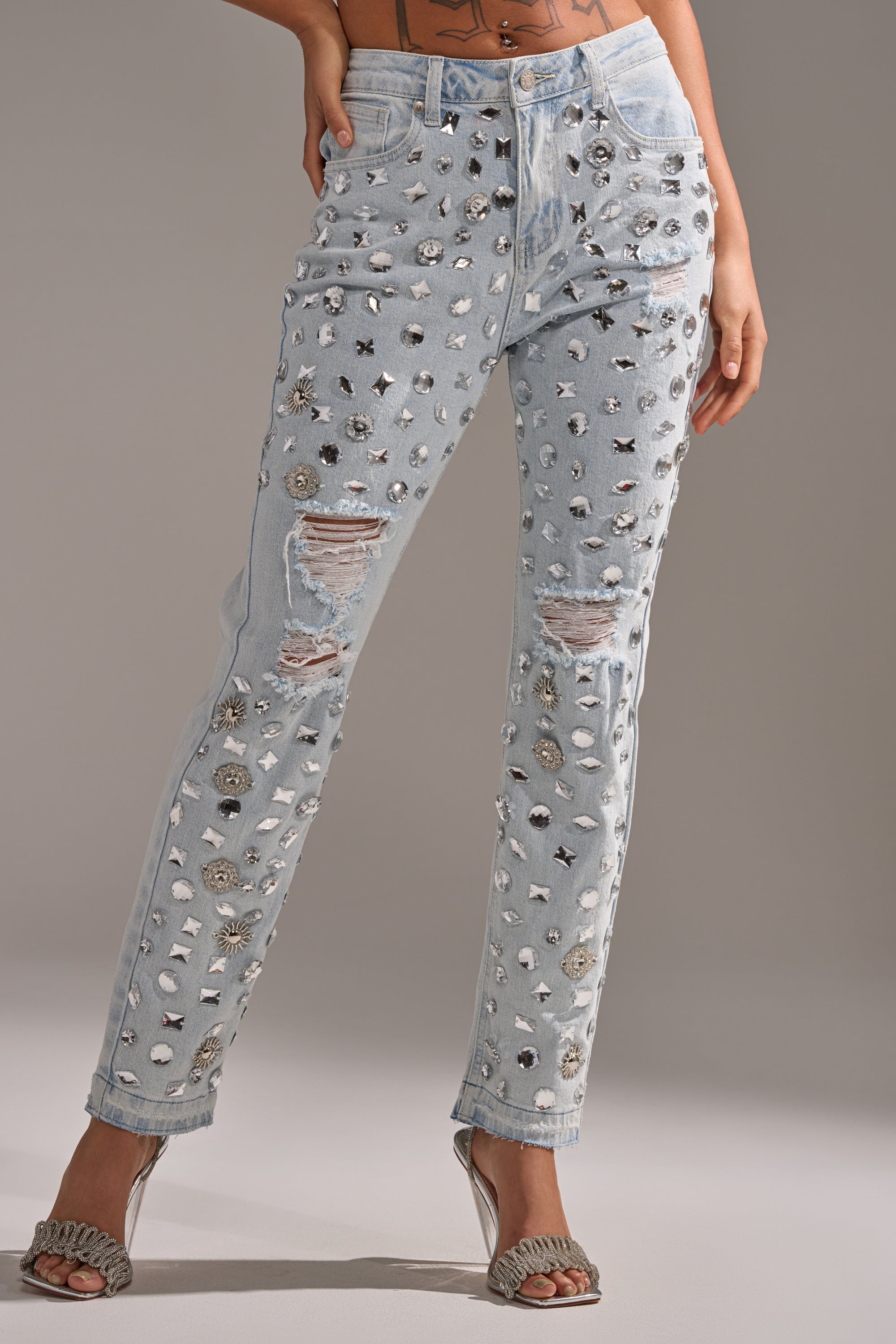 A person wears the DAMN GIRL EMBELLISHED SKINNY DENIM, light blue jeans with metallic embellishments, paired with clear high-heeled sandals against a plain neutral background.
