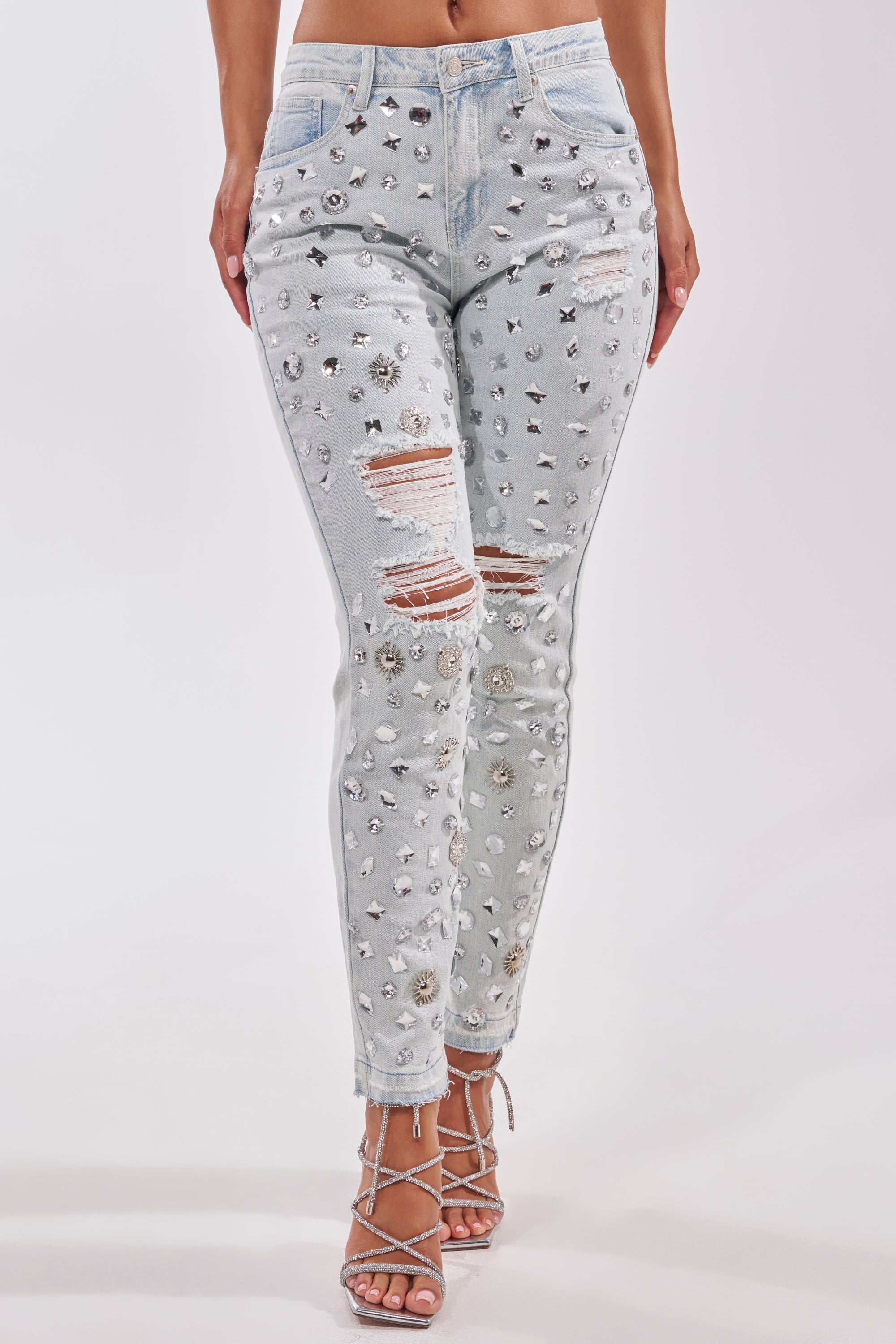 A woman wears DAMN GIRL EMBELLISHED SKINNY DENIM, featuring light-wash, high-waisted, crystal-studded skinny jeans with rhinestone details and strappy heels. The embellished denim pops against a plain white background.