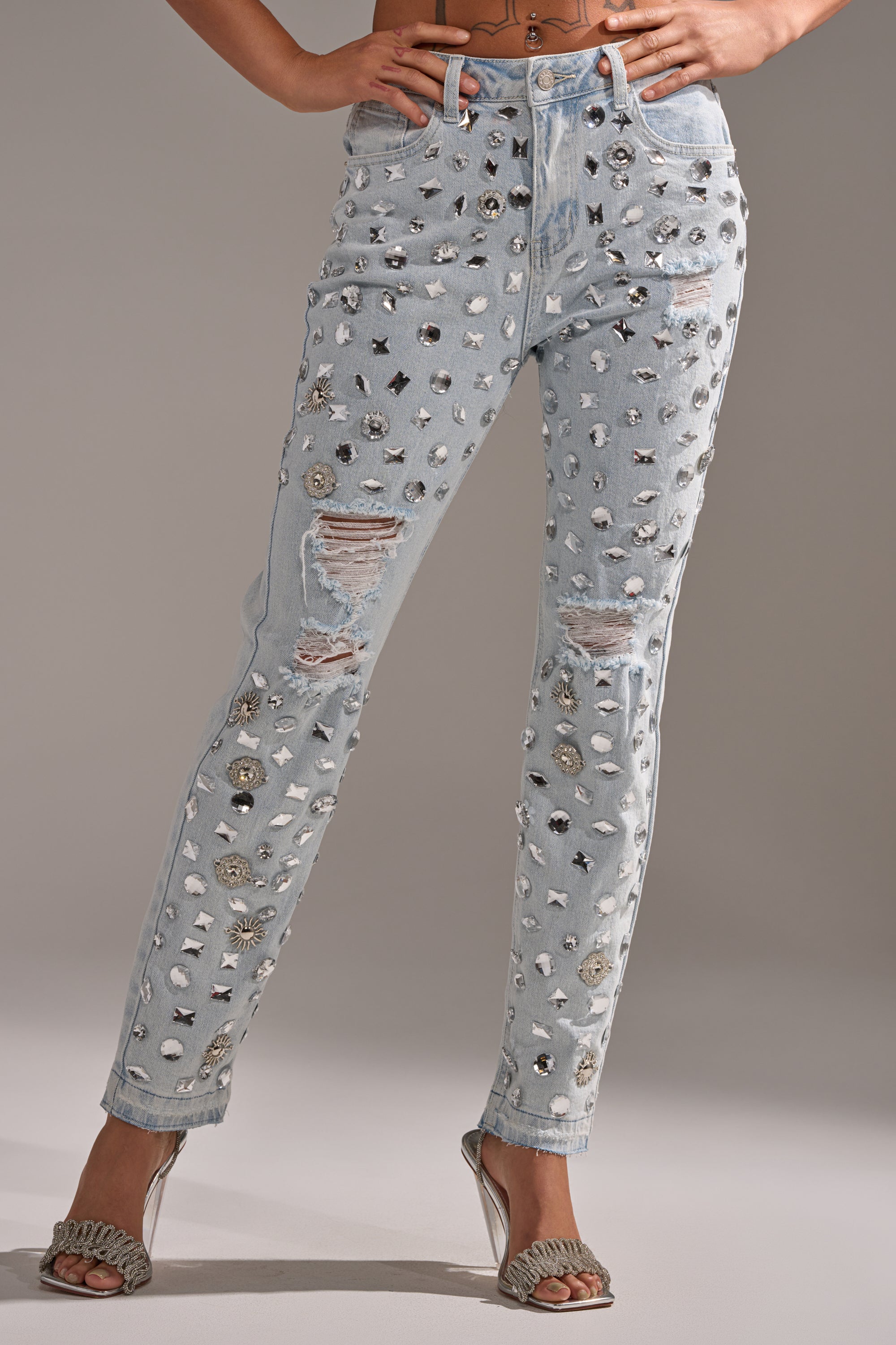 A person wears the DAMN GIRL EMBELLISHED SKINNY DENIM—light blue crystal stud jeans with silver studs and decorative stones—paired with silver high-heeled sandals. The photo is cropped below the chest.