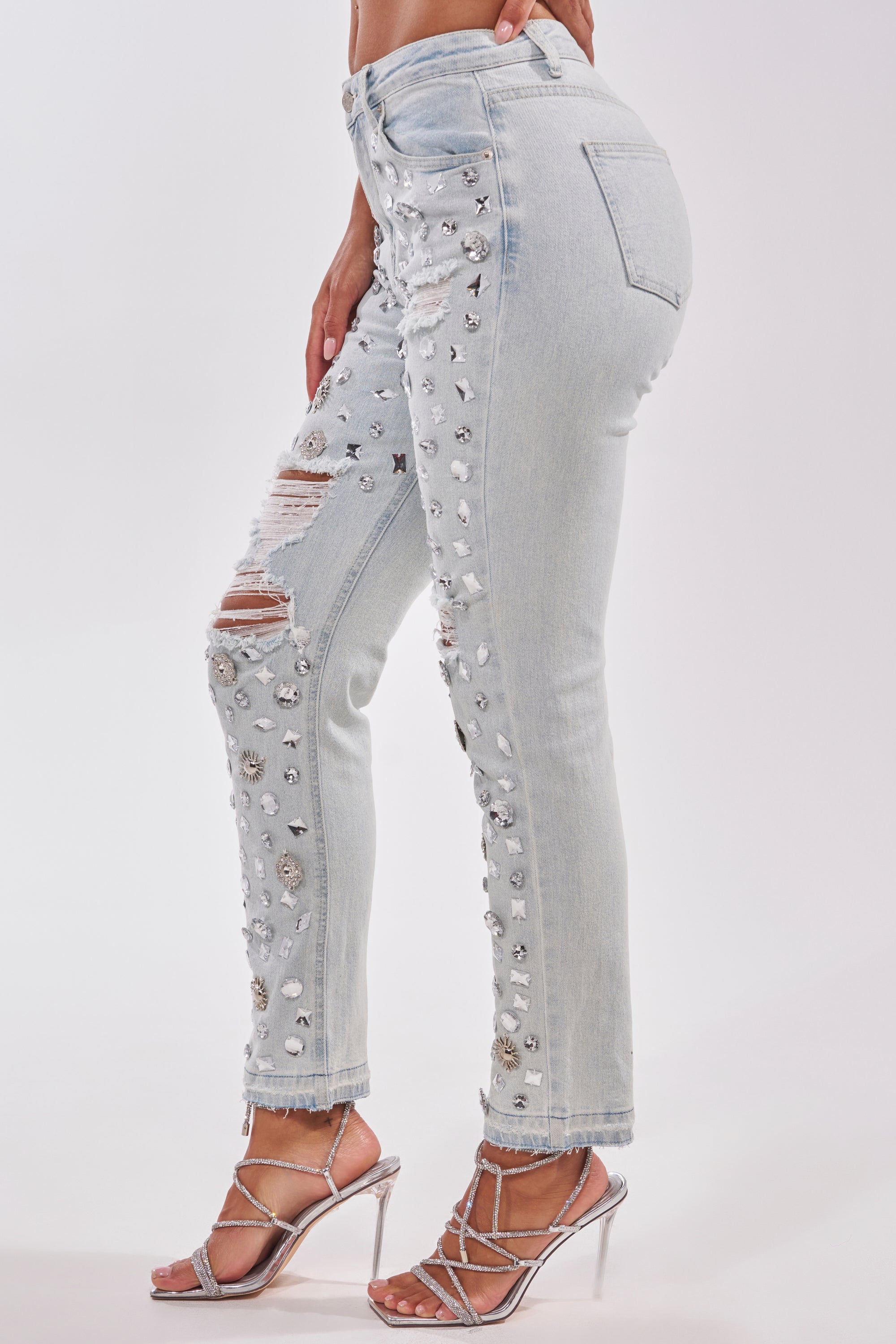 A person wears DAMN GIRL EMBELLISHED SKINNY DENIM, light blue skinny jeans with silver studs and rhinestones, paired with silver strappy high-heeled sandals, standing on a white background.