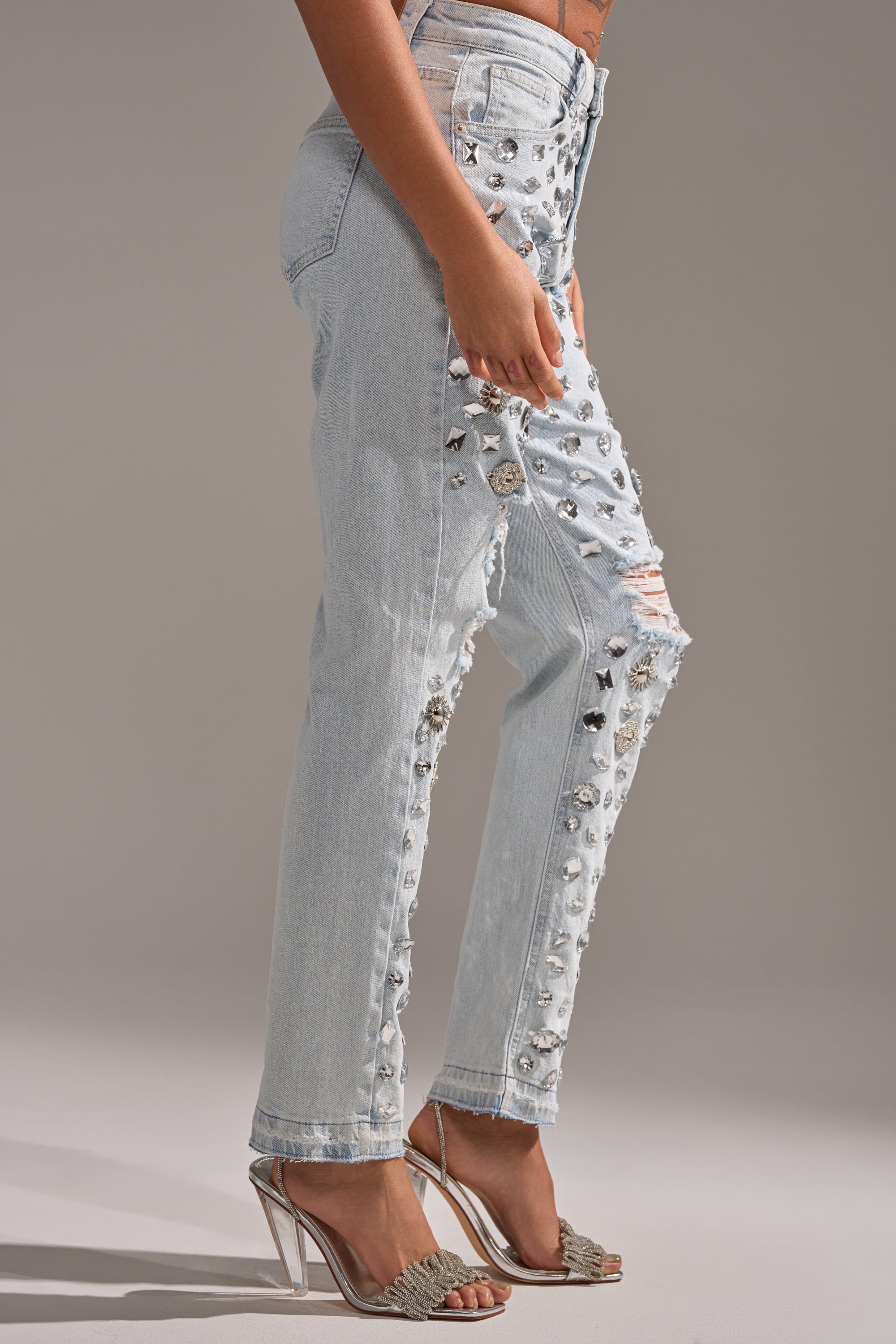 Wearing DAMN GIRL EMBELLISHED SKINNY DENIM—high rise, light wash jeans with rhinestones and rips—and silver high-heeled sandals, a person stands against a plain gray background.