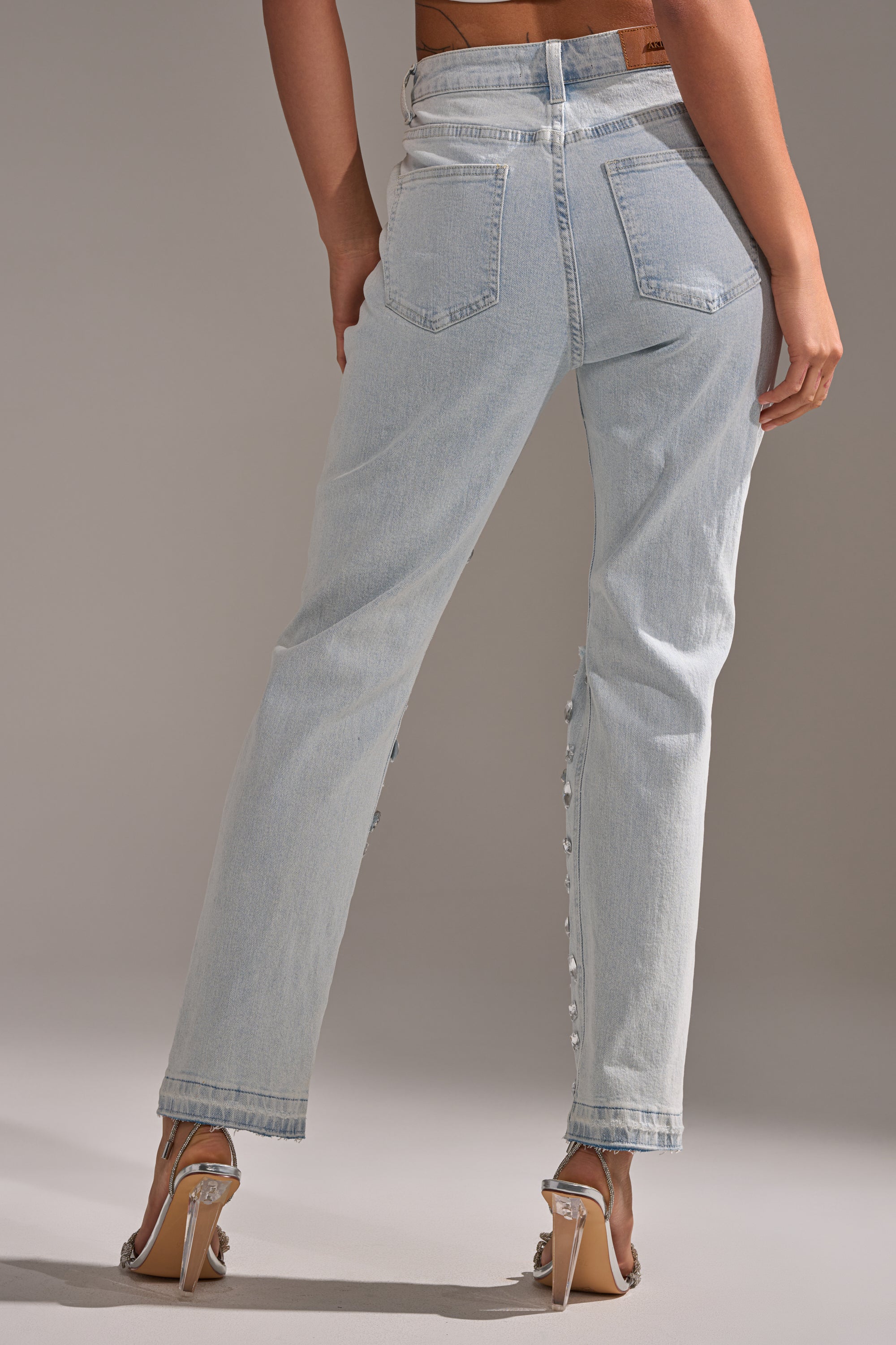 A person stands with their back to the camera, wearing DAMN GIRL EMBELLISHED SKINNY DENIM high-rise stretch jeans with back pockets, side buttons, and silver sandals. The jeans are light blue and feature embellishments.