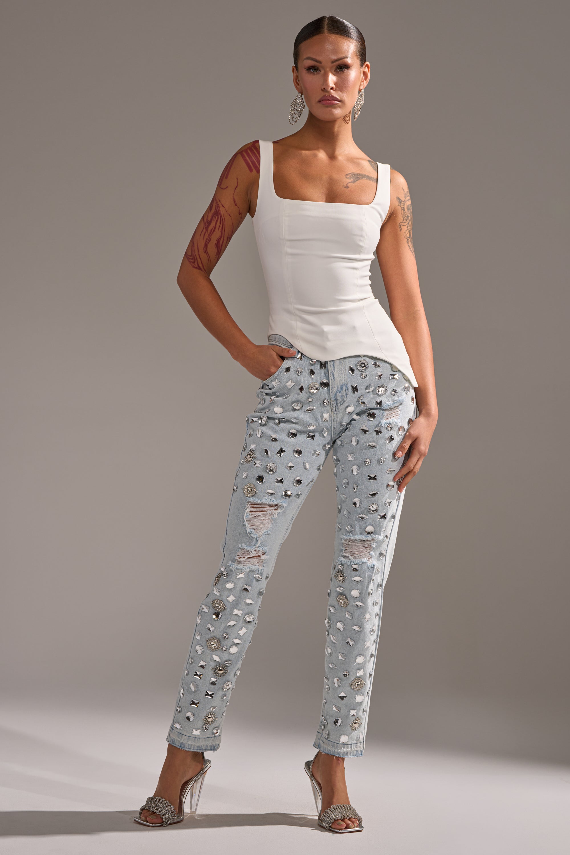 A woman with visible tattoos and hair styled back stands confidently in a fitted white sleeveless top, DAMN GIRL EMBELLISHED SKINNY DENIM jeans, and silver high-heeled sandals.