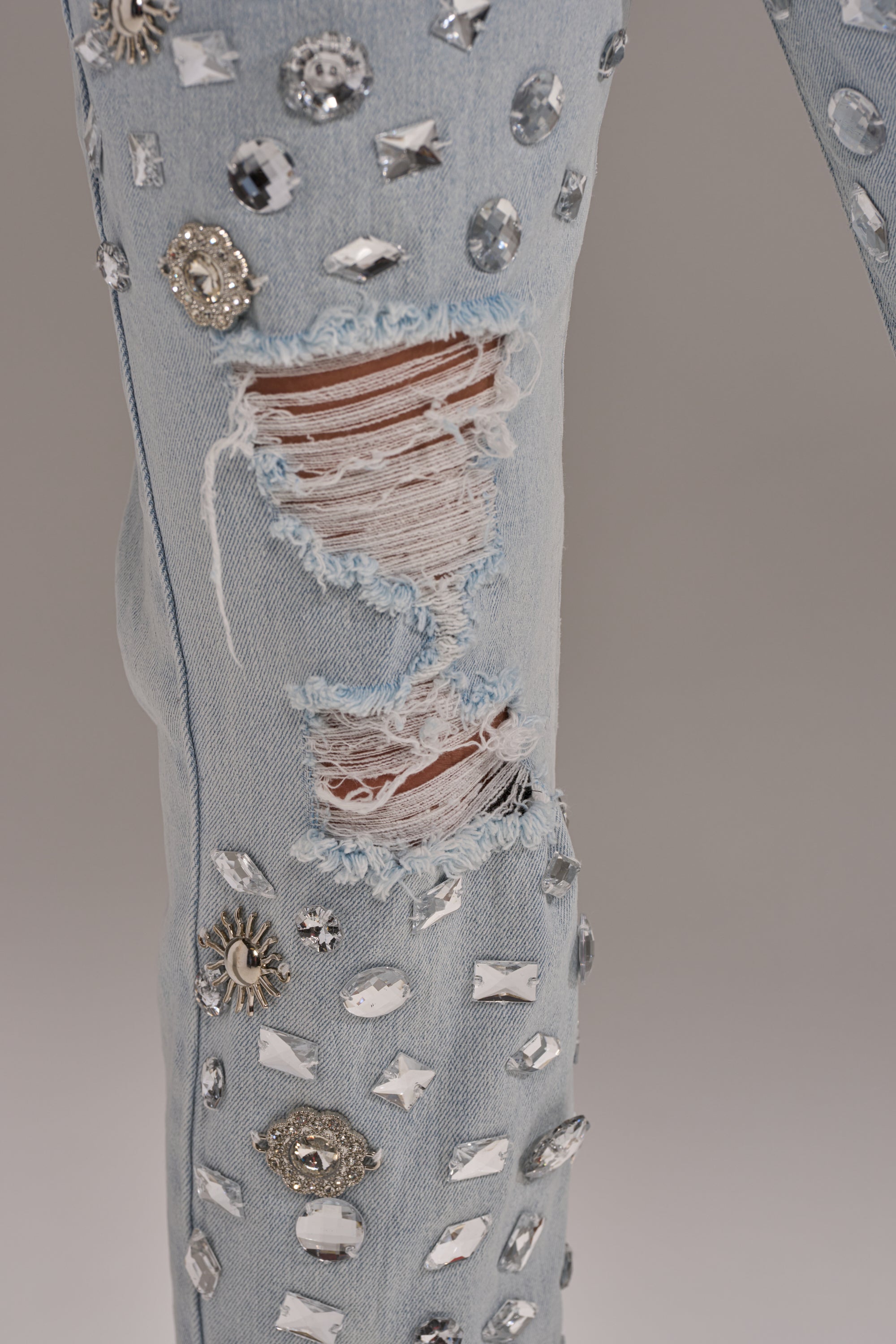 Close-up of DAMN GIRL EMBELLISHED SKINNY DENIM, a high-rise light wash jean with a large ripped section revealing threads and skin, adorned with silver and clear rhinestones and metal accents for a sparkling crystal-studded look.