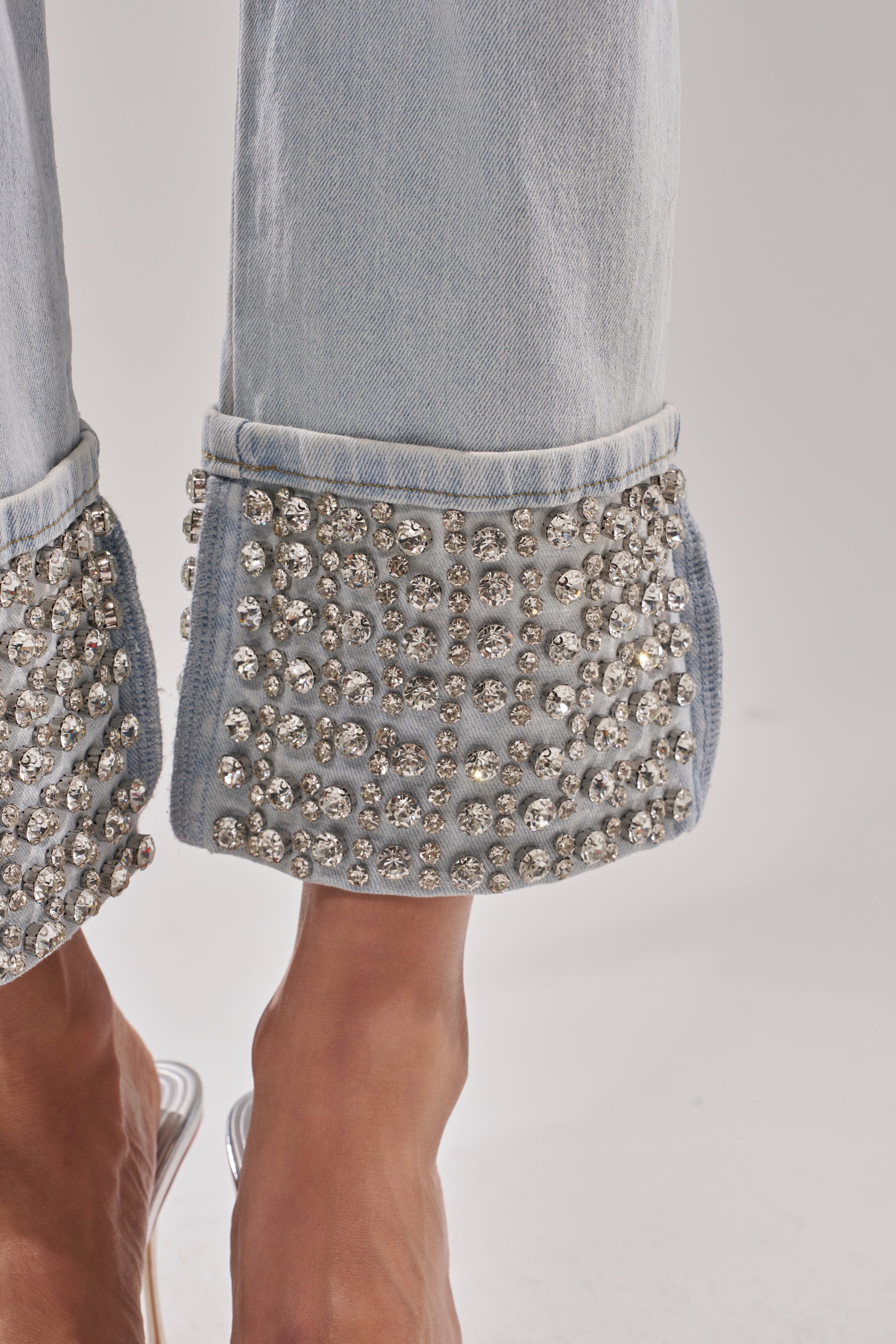 Close-up of the HANNI RHINESTONE DENIM CROPPED PANT in a high-rise fit, featuring cuffs embellished with large sparkling rhinestones. Bare feet and clear-heeled shoes appear against a plain neutral backdrop. 9