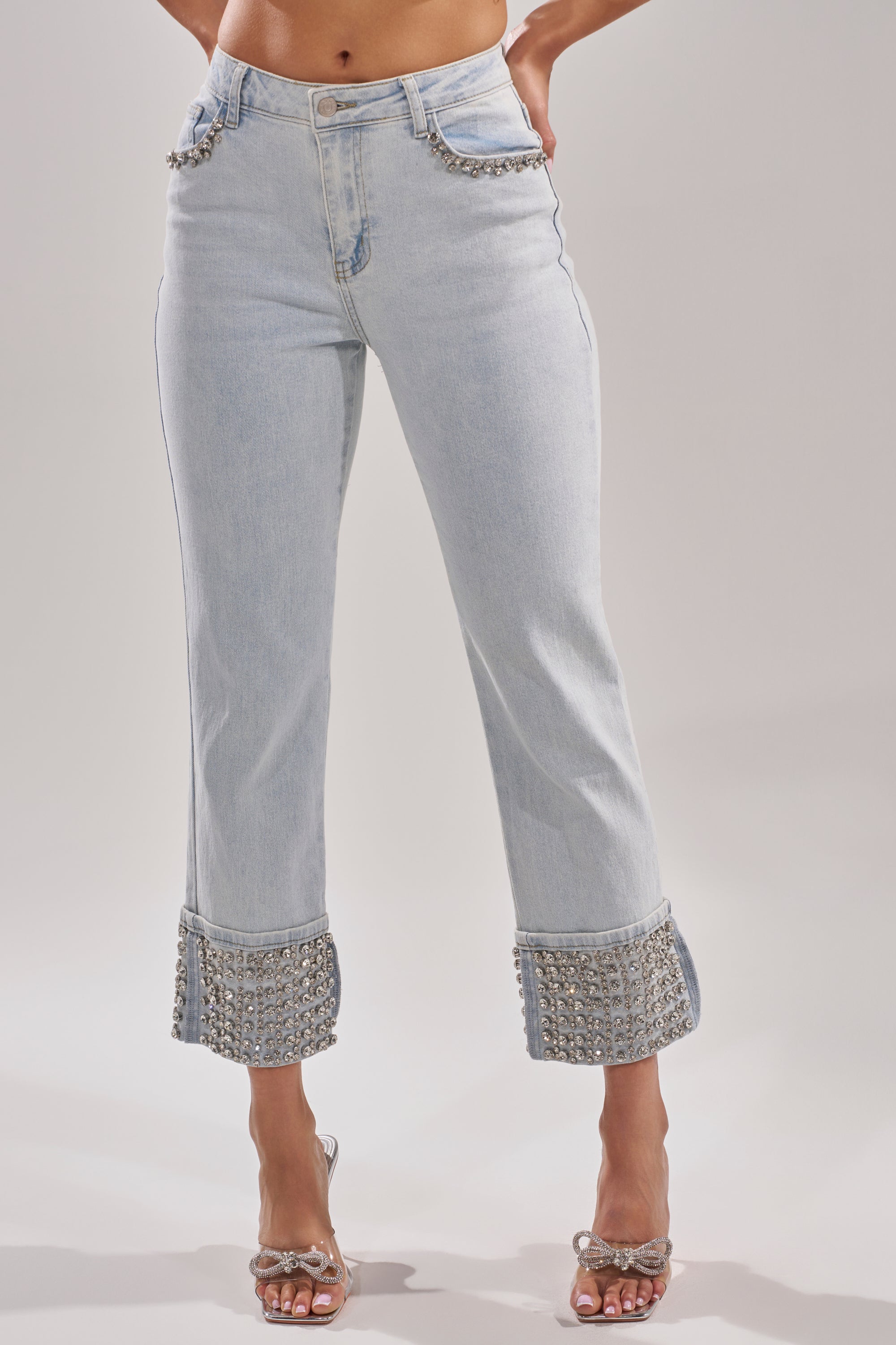 Someone wears the HANNI RHINESTONE DENIM CROPPED PANT, featuring high-rise, light wash denim with pearl details on pockets and cuffs, paired with transparent heeled sandals. The upper body is not shown. 1