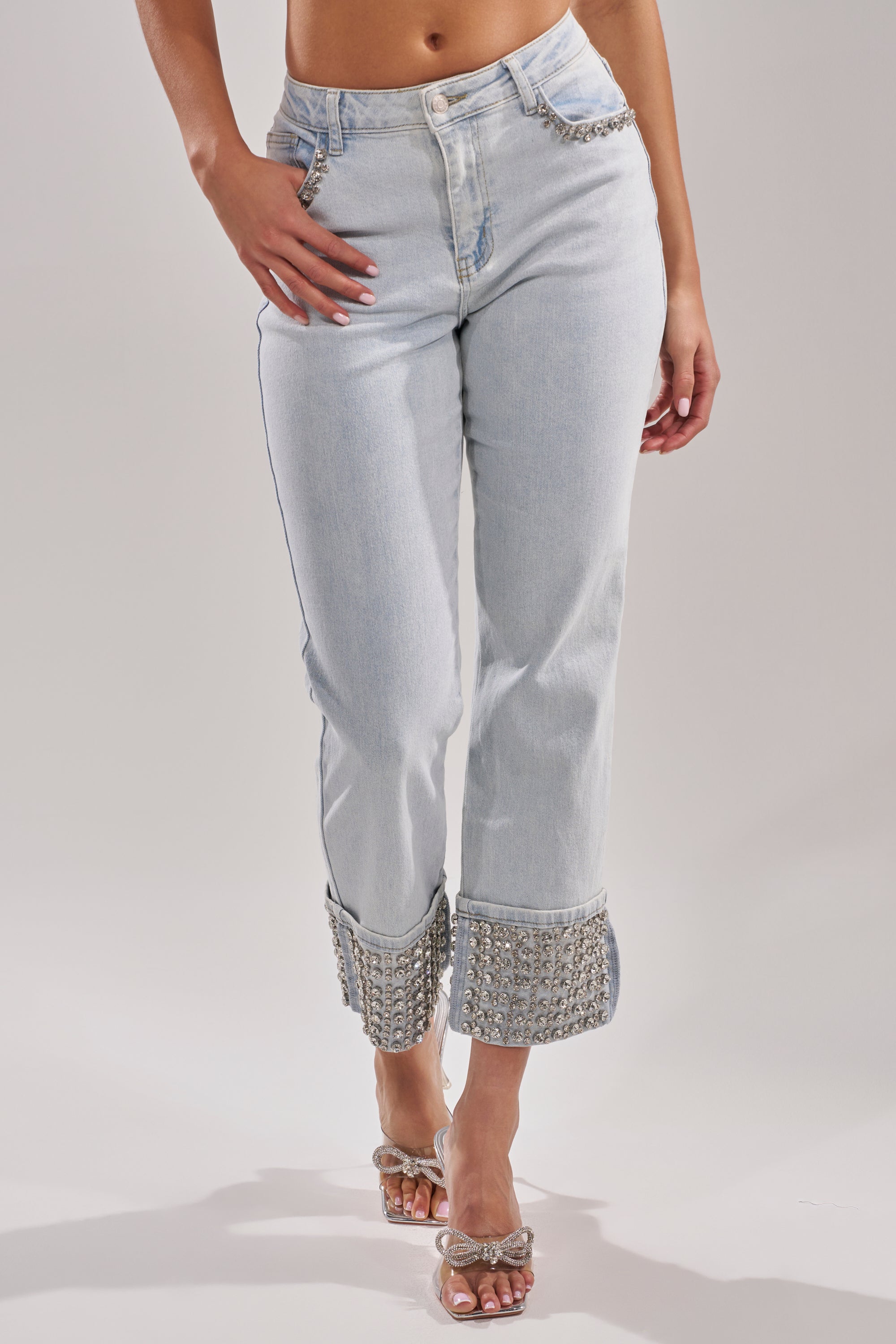 A model wears the HANNI RHINESTONE DENIM CROPPED PANT, featuring a light-wash, high-waisted fit with dazzling chain pocket accents and sparkling cuff embellishments, styled with transparent crystal-detailed heels. 2